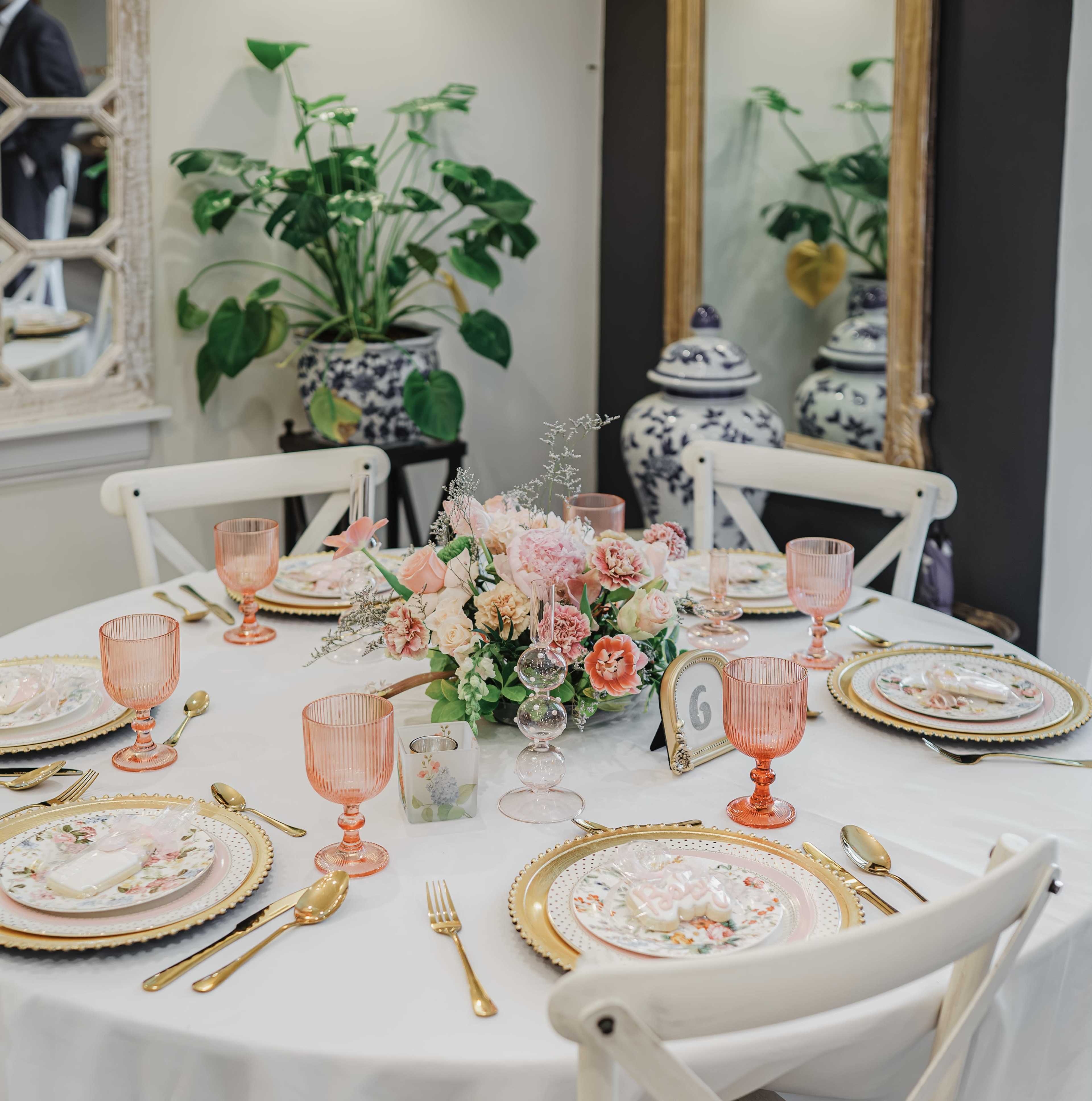 A elegantly set dining table with pastel-colored tableware, a floral centerpiece, and decorative glassware, surrounded by indoor plants and mirrors.