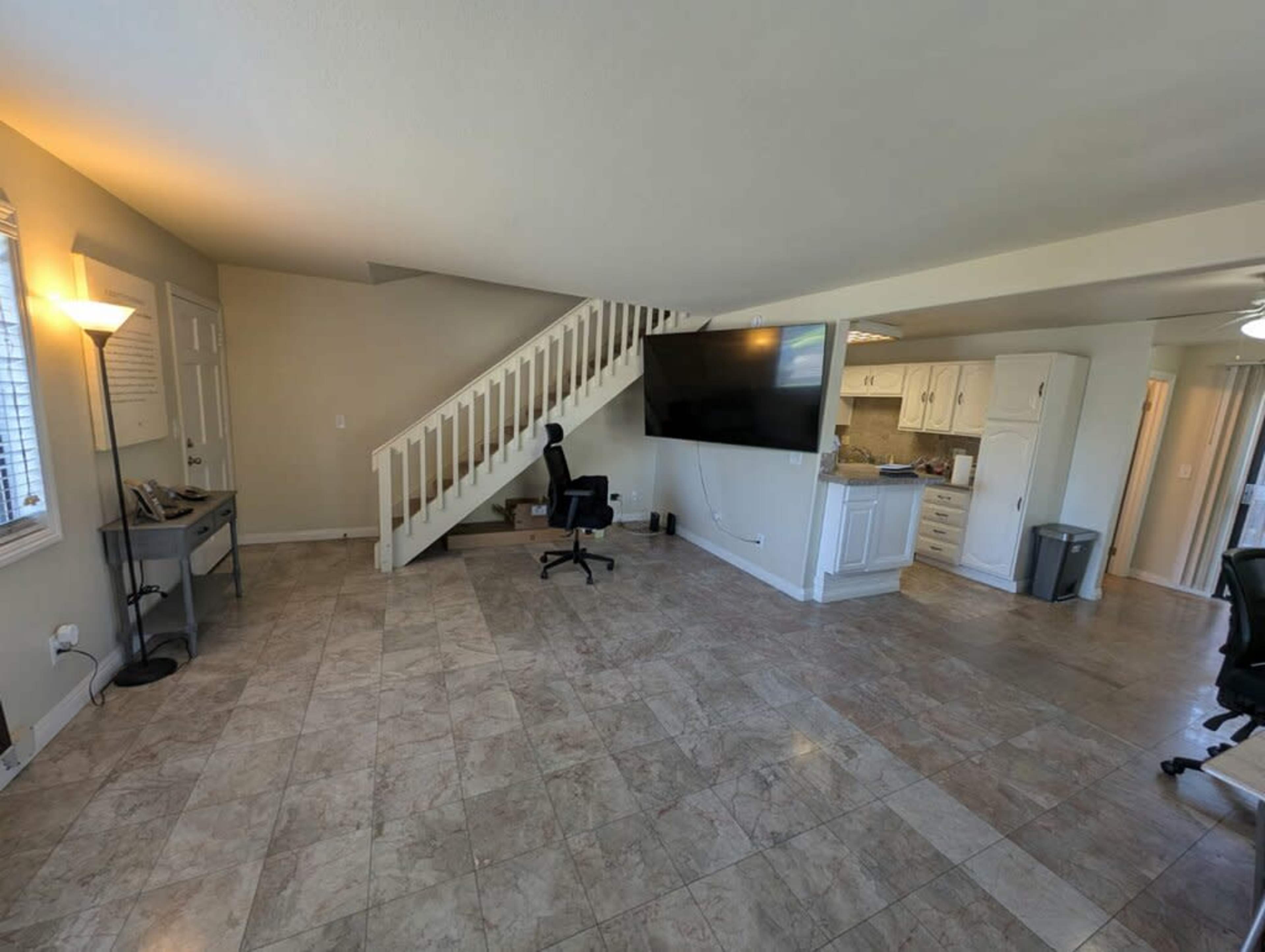 The image shows a spacious room with tiled flooring, a staircase on the left, a large television mounted on the wall, and several office chairs positioned around the space.