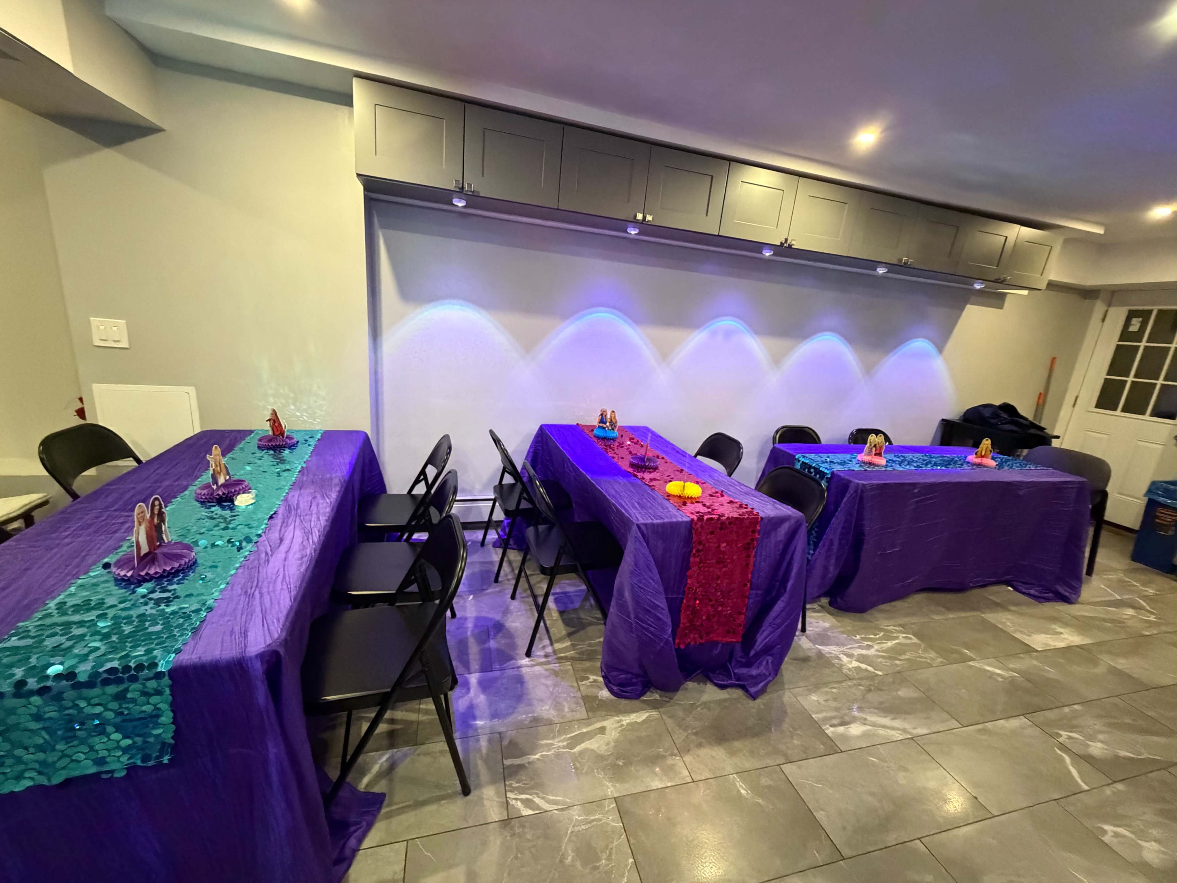 The image shows a room with four tables covered in purple and red sequined tablecloths, arranged with decorative items on top, under ambient lighting.