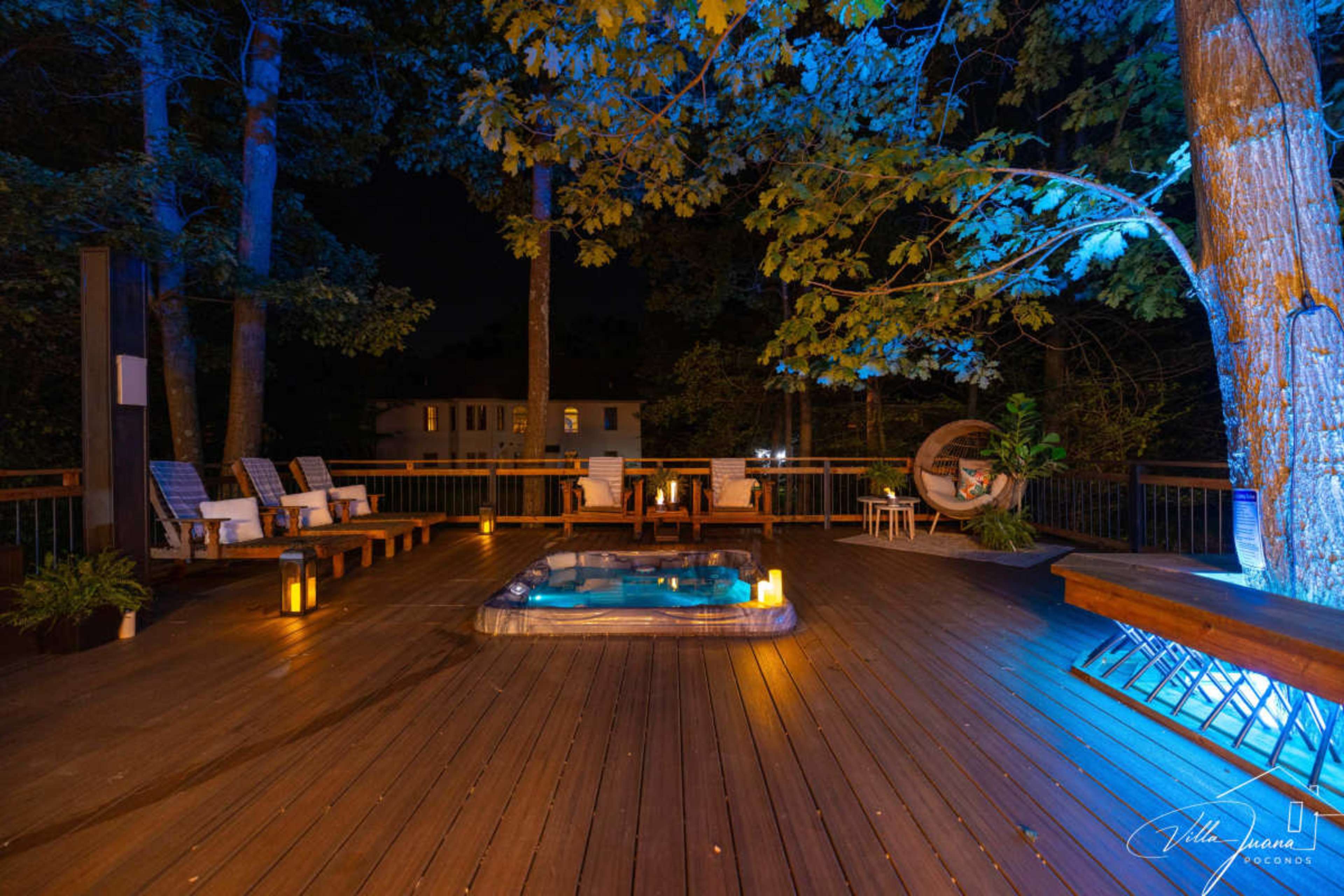 A wooden deck features a hot tub surrounded by lounge chairs and lanterns, illuminated by blue lights at night.