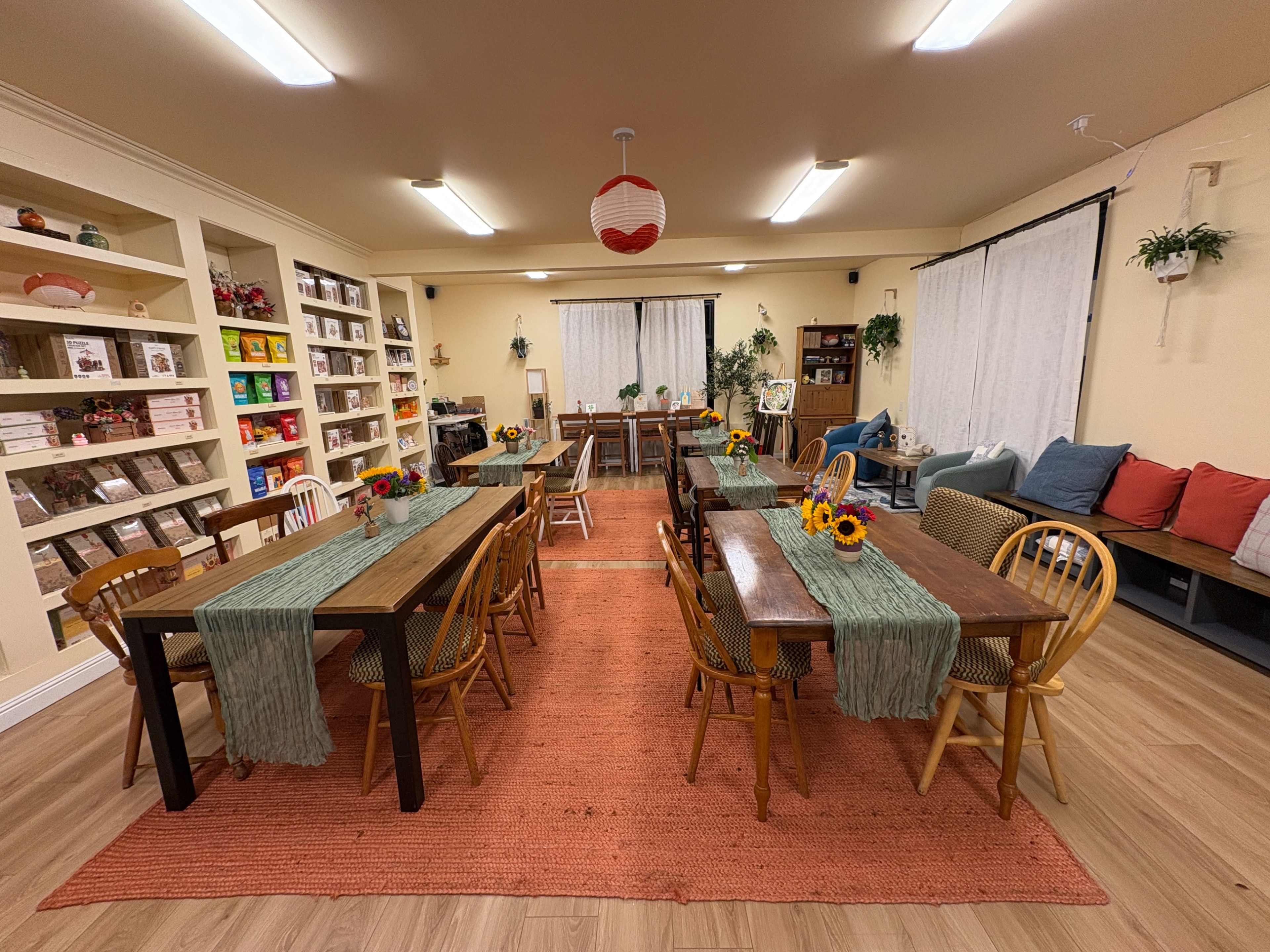 The image shows a spacious room set up with several wooden tables and chairs arranged for group activities, decorated with flowers and a large rug, surrounded by shelves filled with various items.