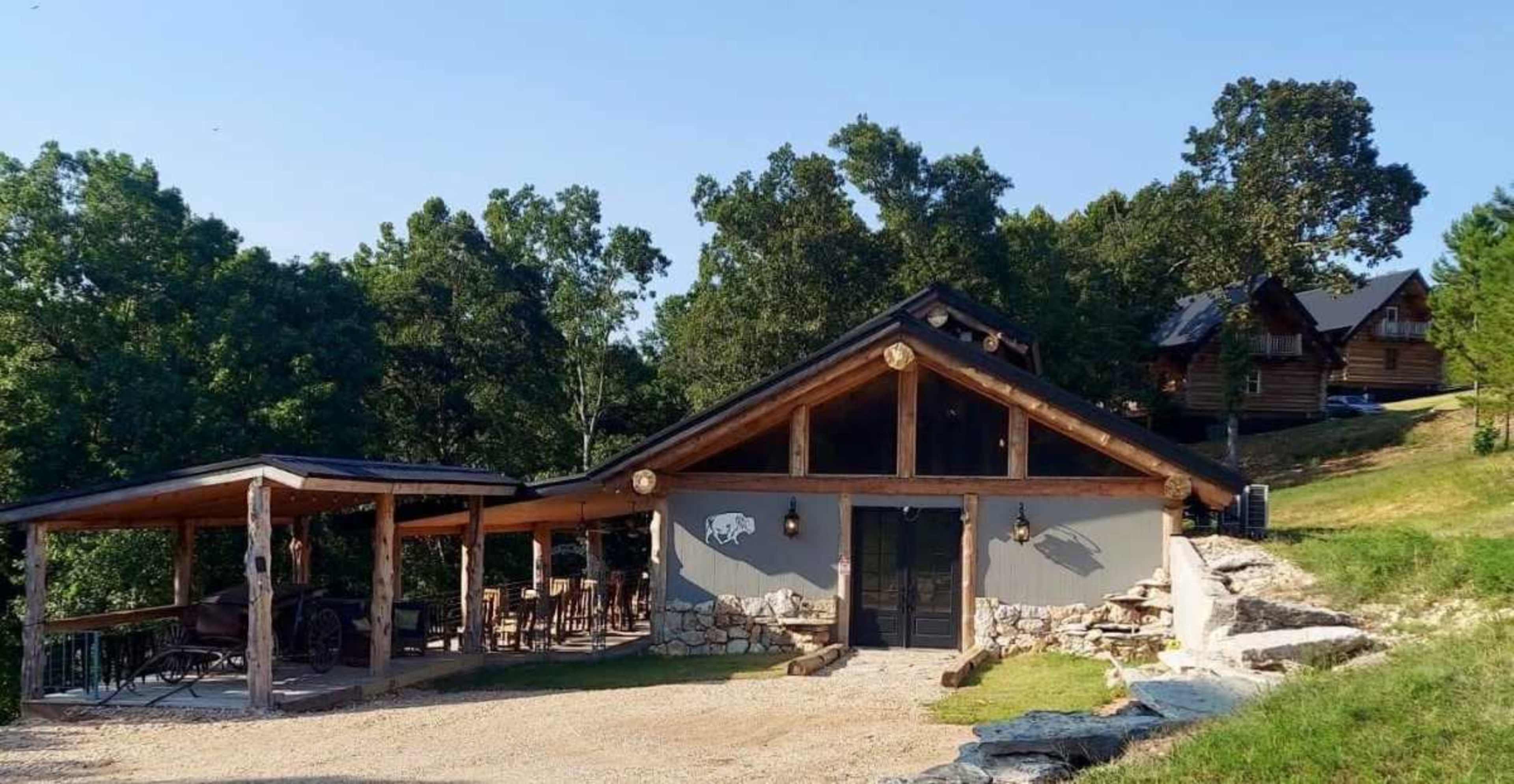 The image shows a rustic cabin with a stone and wood exterior, surrounded by trees and featuring an outdoor seating area.