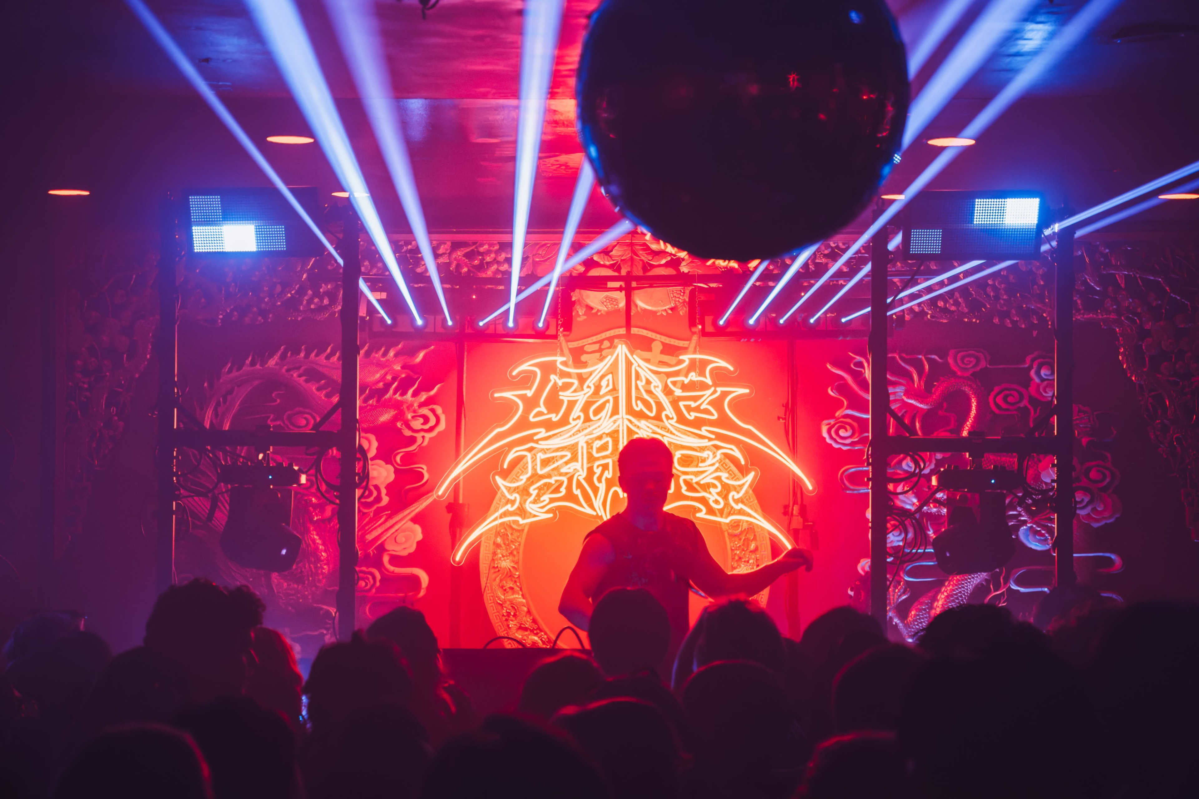A performer stands on stage in front of a vibrant neon backdrop while colorful lights illuminate a crowded venue.