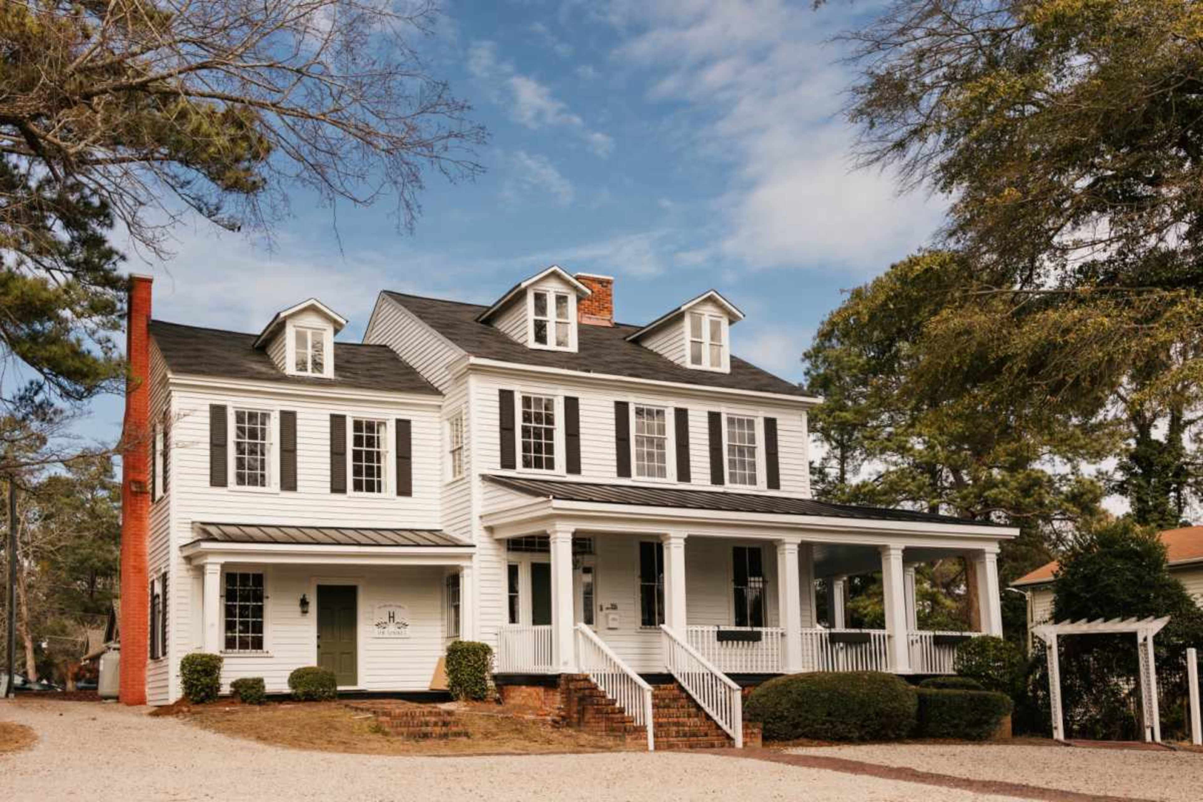 The image shows a large white two-story house with a front porch, black shutters, and a combination of brick and siding, surrounded by trees and a gravel driveway.