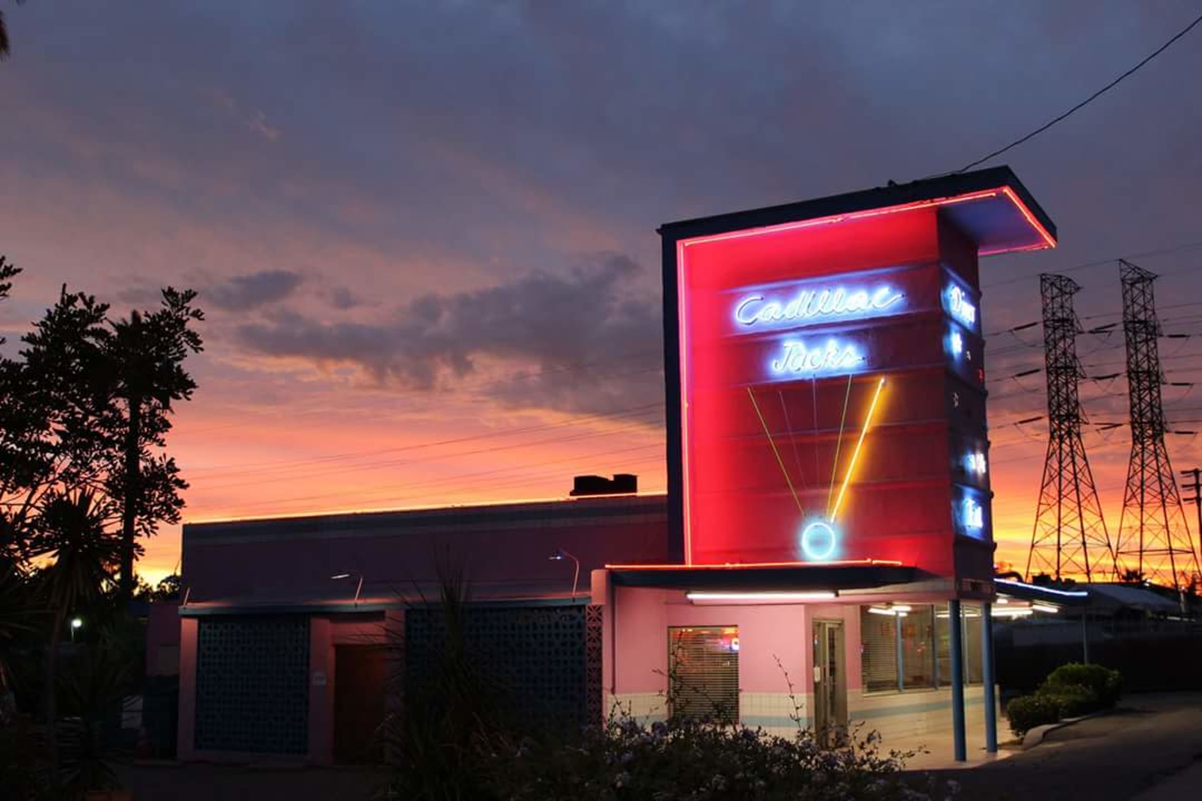 A neon-lit building with a "Cadillac Jack's" sign stands against a colorful sunset sky, surrounded by power lines and trees.