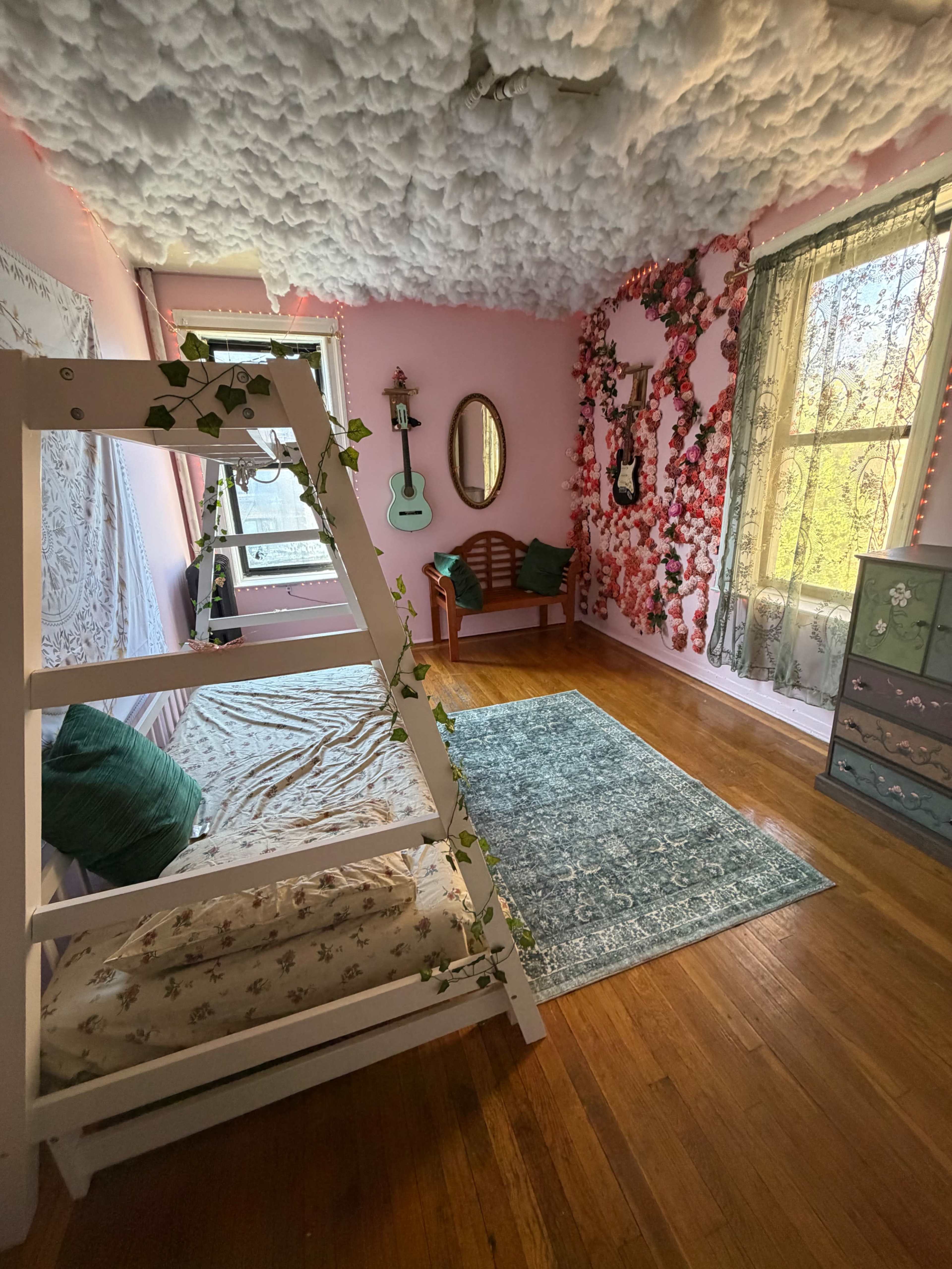The image shows a pastel pink bedroom with a loft bed, decorative floral wall, a green rug, and a small seating area containing a wooden bench and mirror.