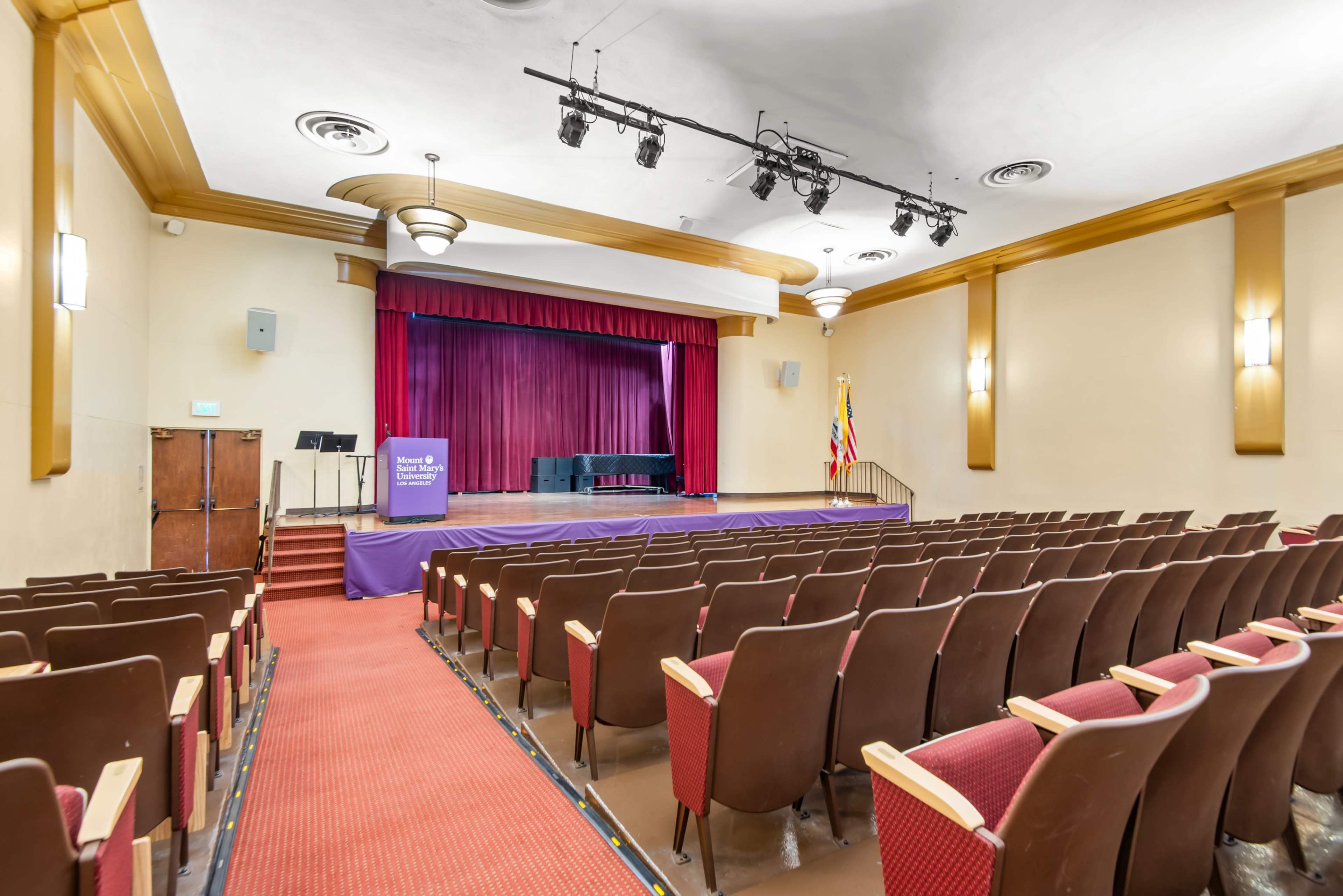 The image shows an empty auditorium with a stage featuring a maroon curtain and seating arranged in rows.