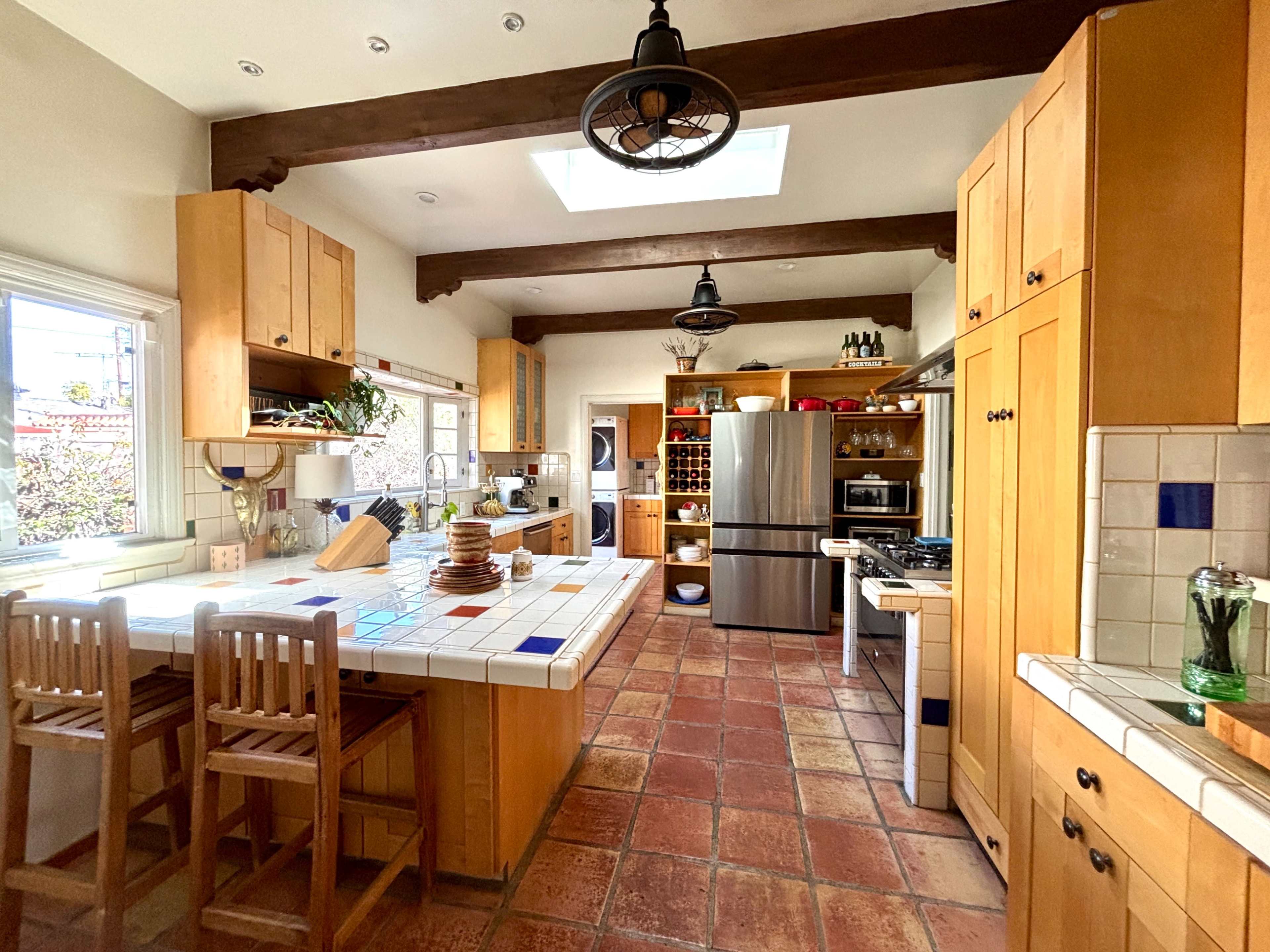 The kitchen features wooden cabinets, a central island with bar stools, stainless steel appliances, and a skylight for natural light.