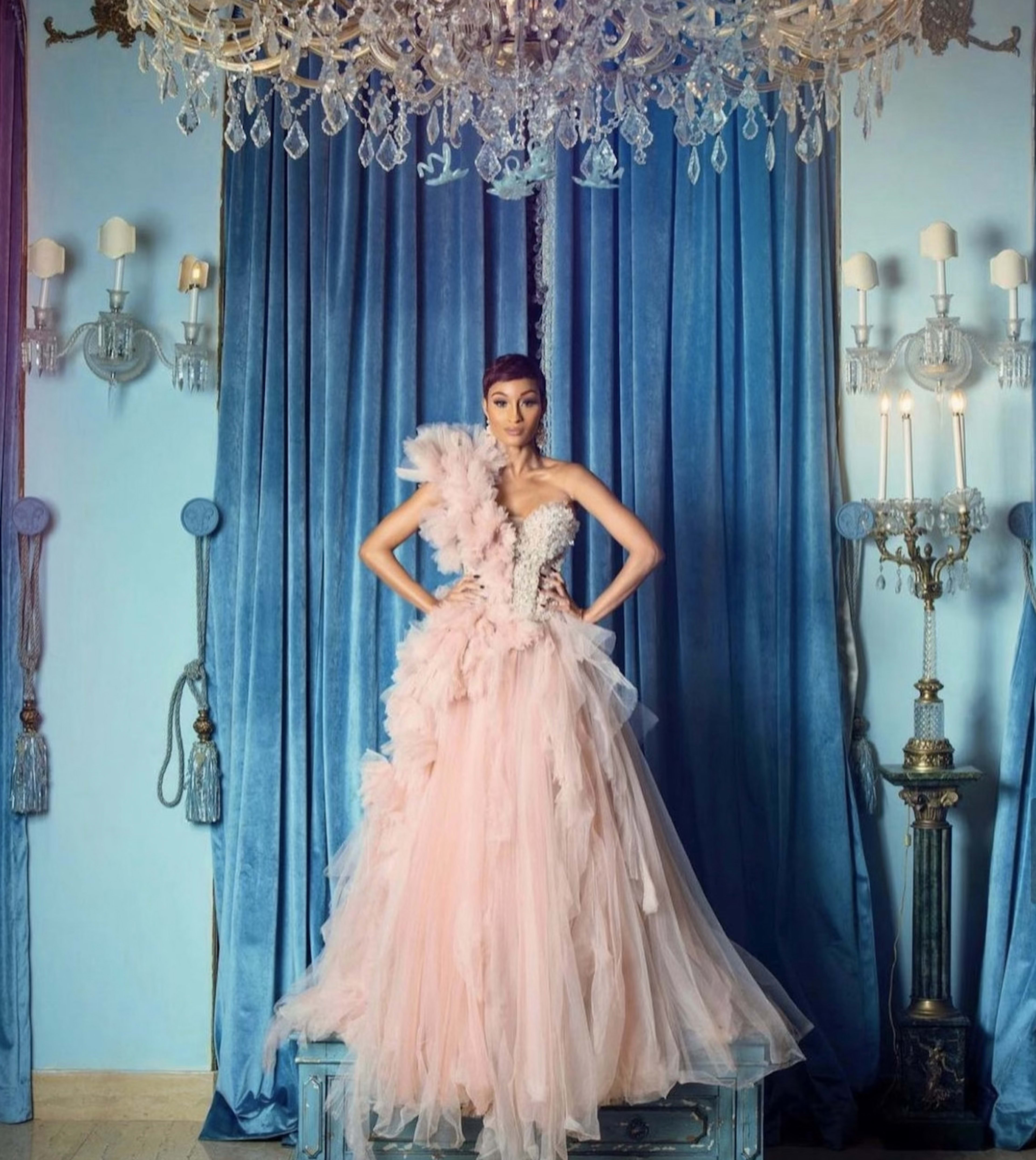 A model poses in an elaborate pink gown with feathers in front of a blue backdrop and ornate decor, including a chandelier and wall sconces.