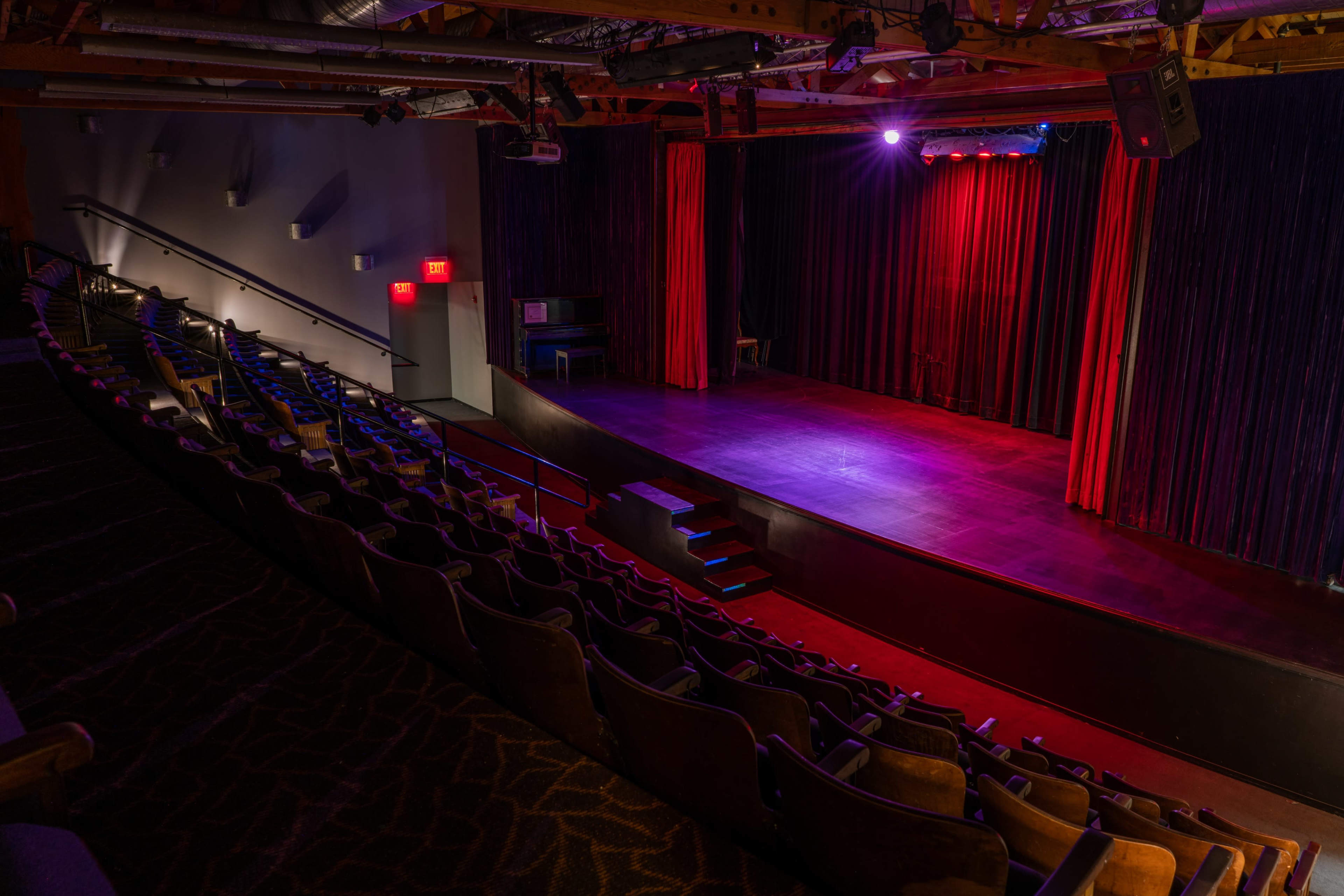 An empty theater with rows of seats facing a small stage illuminated by red and purple lights.