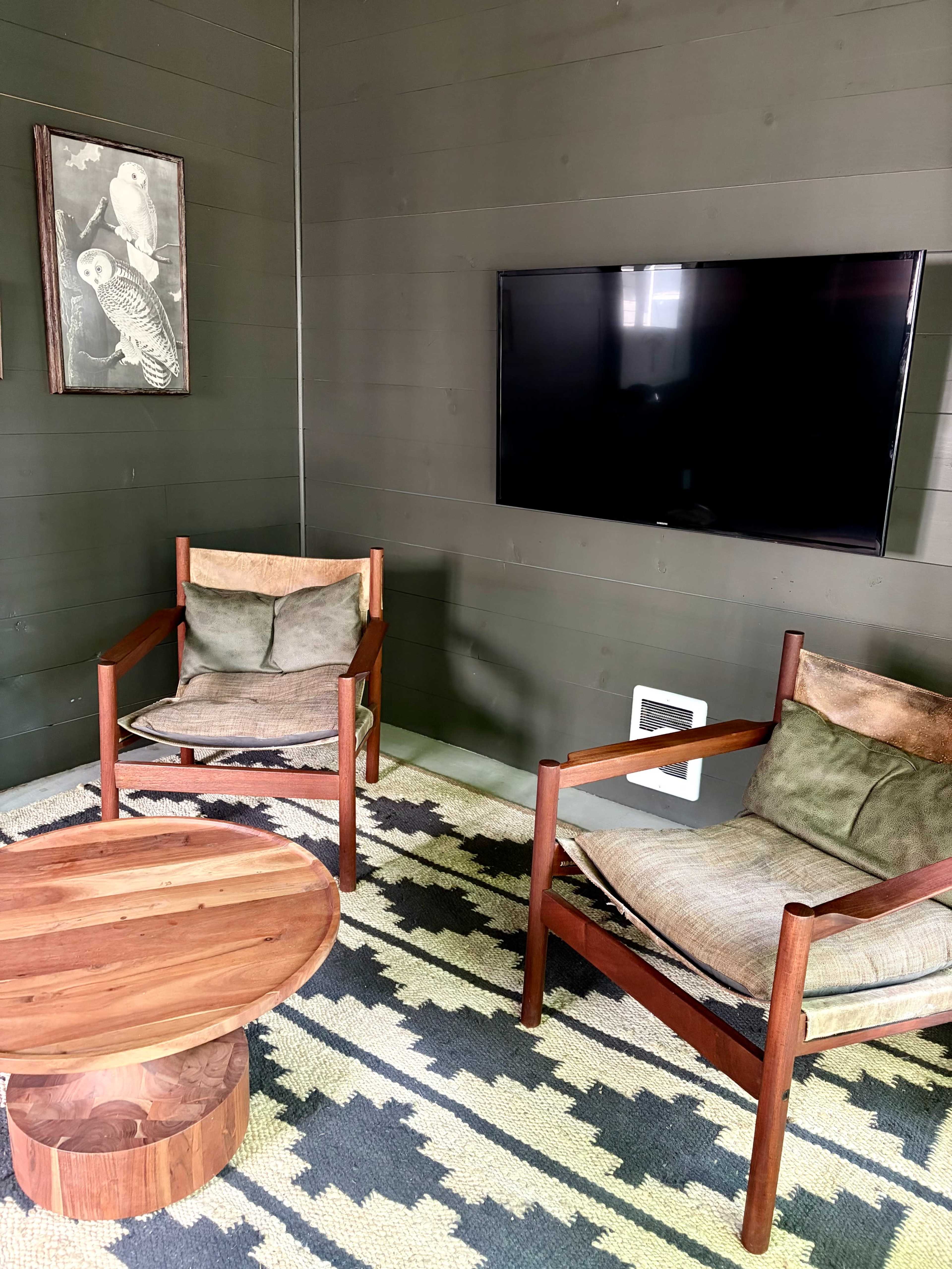 The image shows a cozy sitting area with two wooden chairs, a round wooden coffee table, and a flat-screen television mounted on a dark green wall.