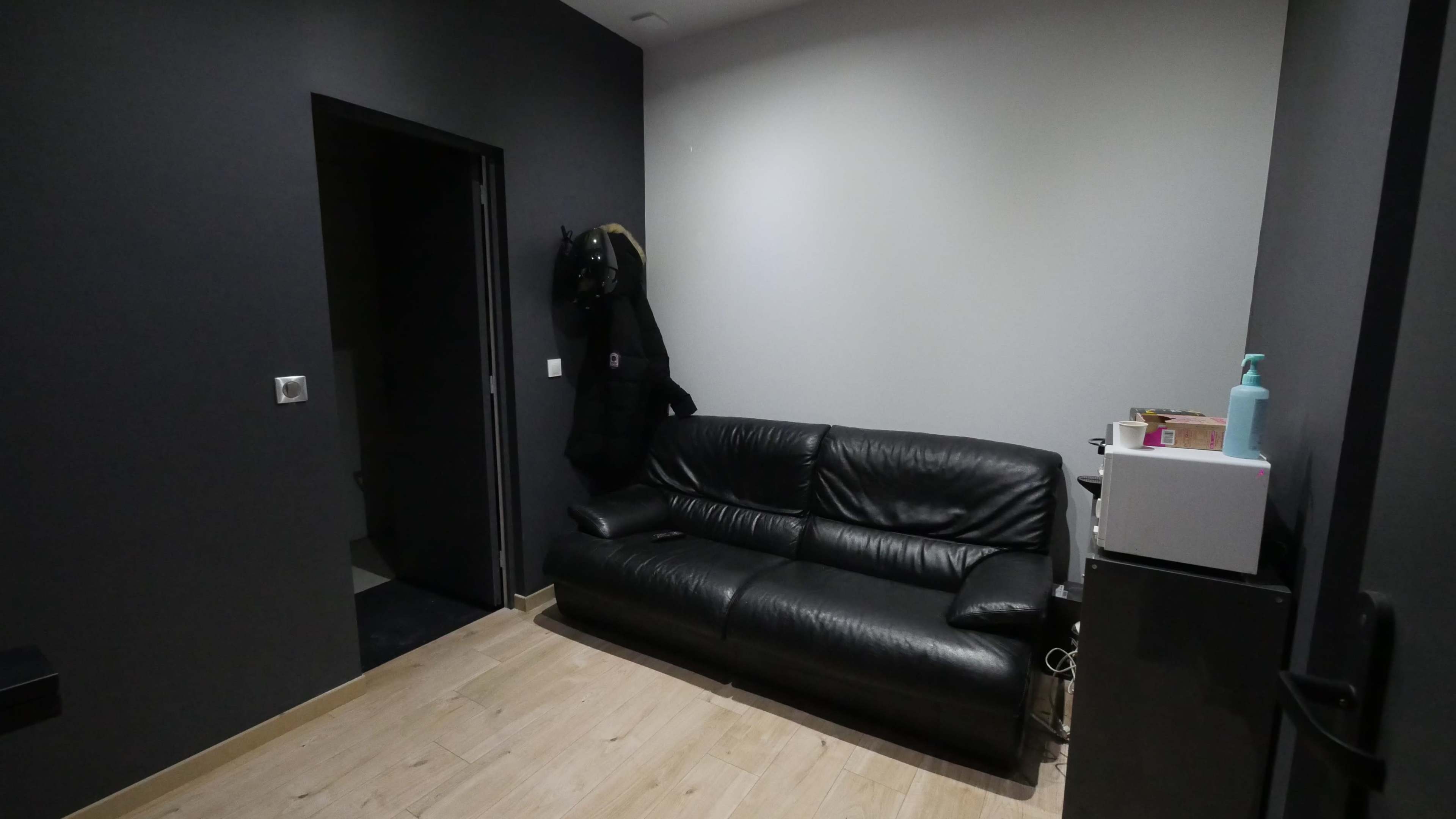 A small room with a black leather couch, a doorway leading to another space, and a wall-mounted coat hanger with a black coat.