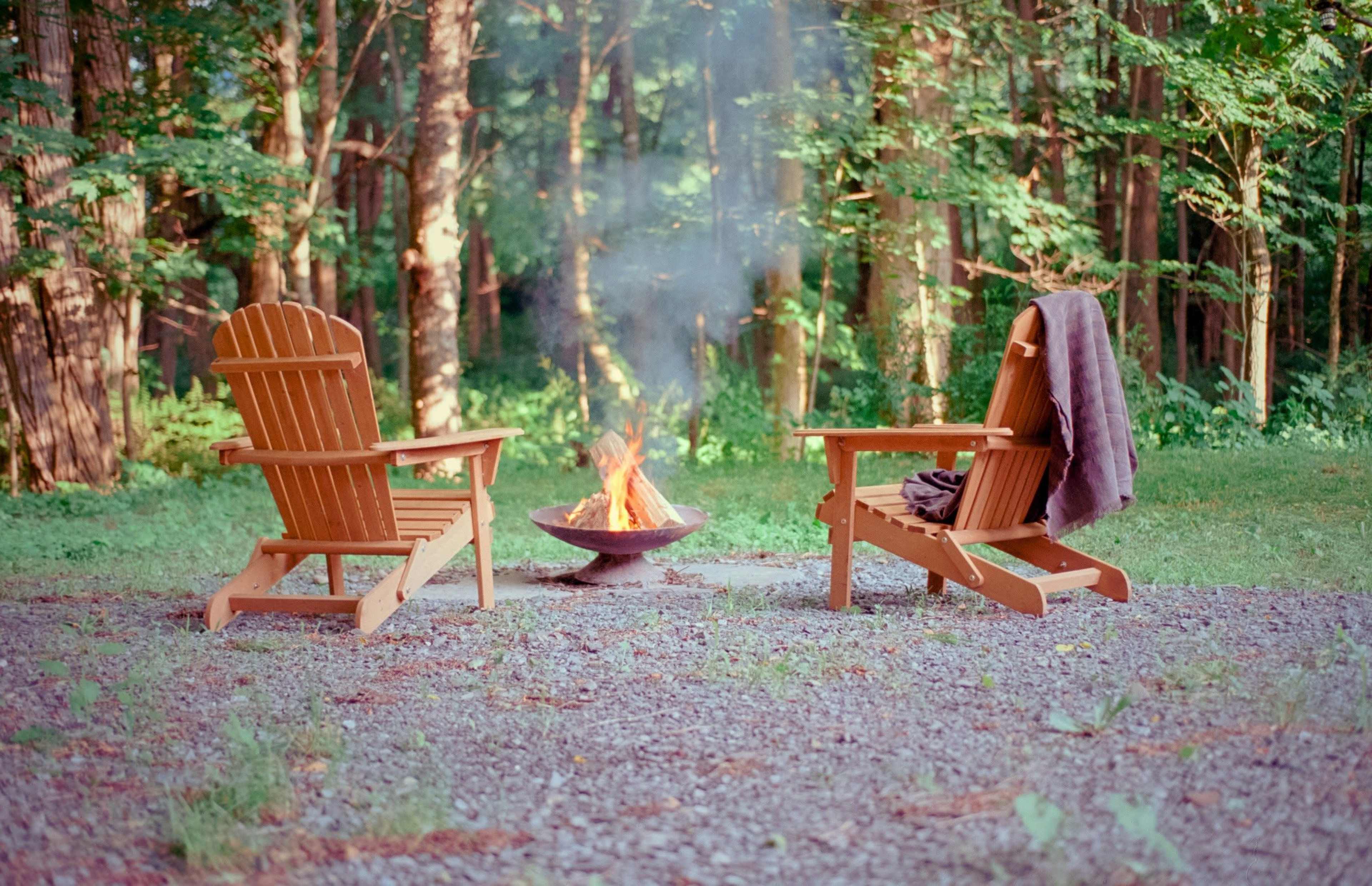 Two wooden Adirondack chairs face a fire pit with an open flame in a forest clearing surrounded by trees.