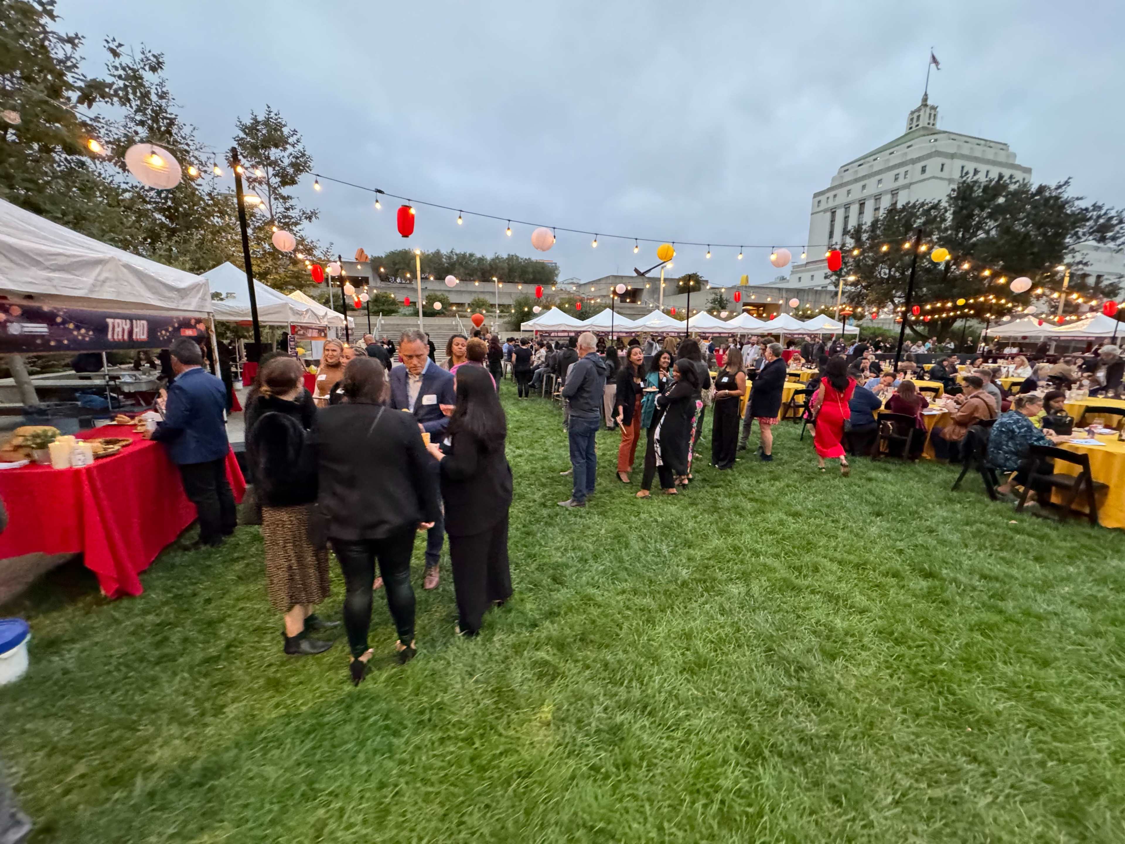 A crowd of people gathers around food stalls and tables, illuminated by string lights and lanterns, on a grassy area near a large building during an outdoor event.