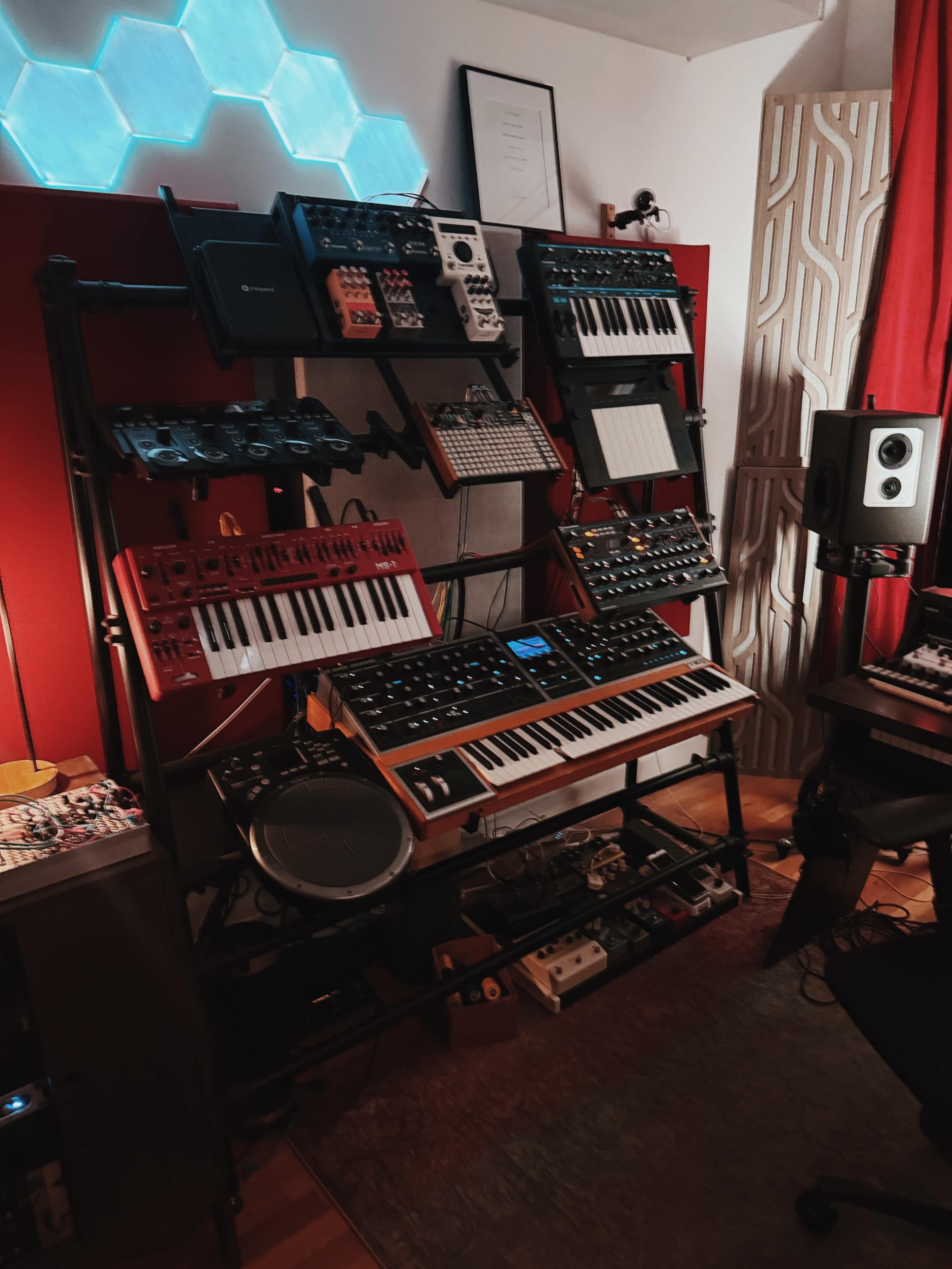 The image shows a music production setup featuring various synthesizers, MIDI controllers, and effects units arranged on a rack against a red wall.