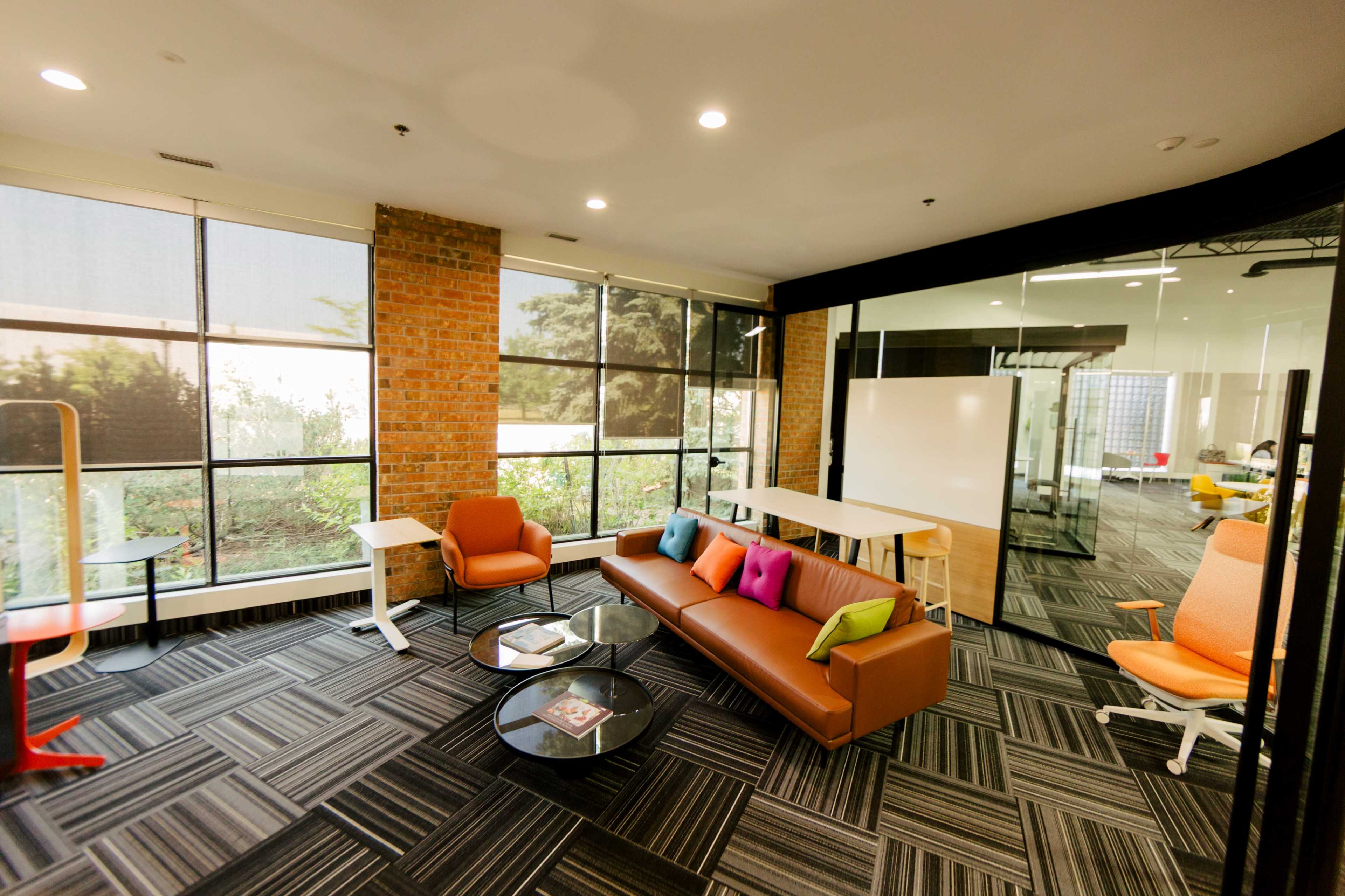 The image shows a modern office space with large windows, a brown couch with colorful cushions, and a glass partition separating it from a conference area.