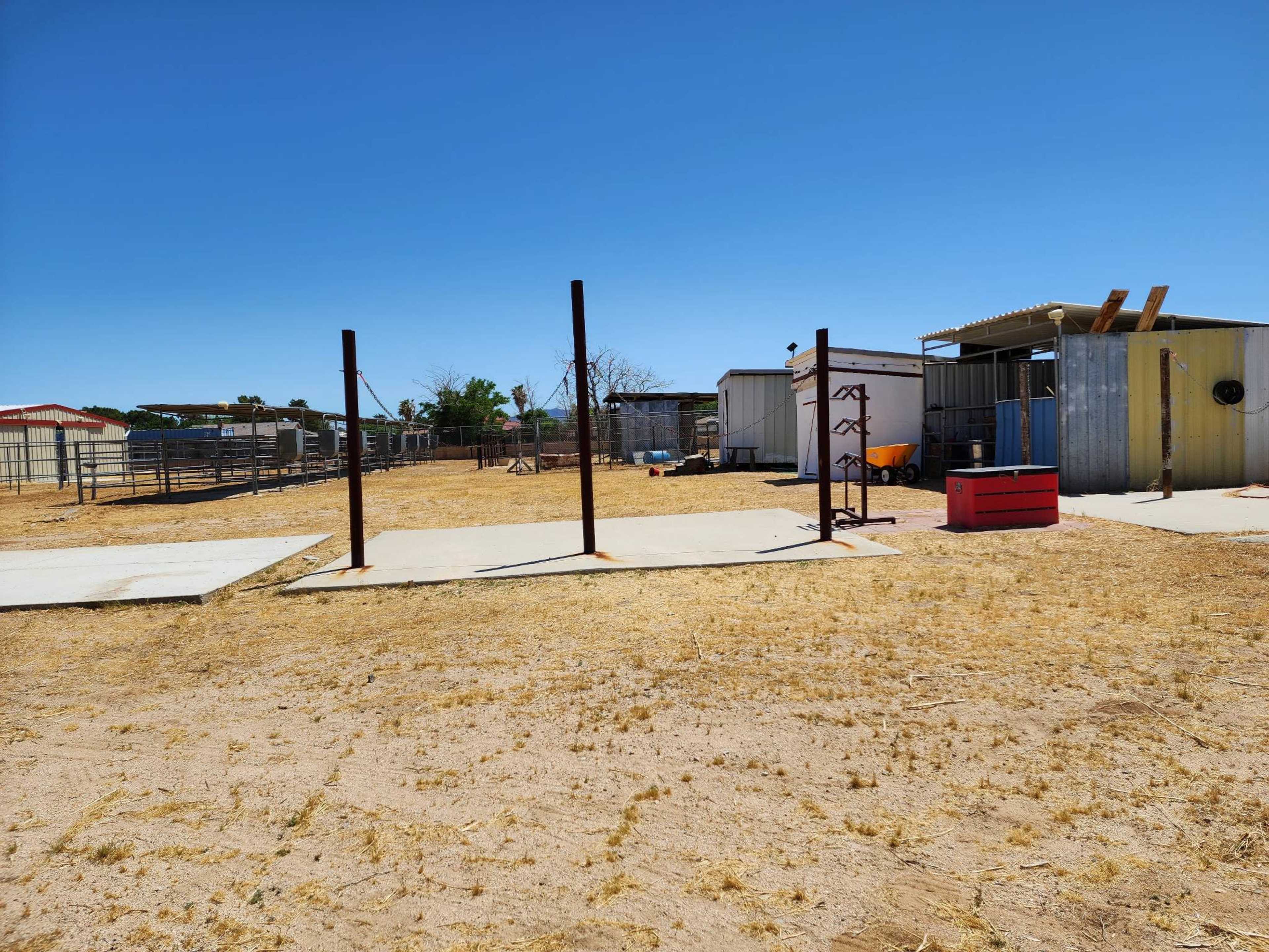 The image shows an outdoor area with several concrete pads, two steel posts for equipment, and a few storage sheds under a clear blue sky.