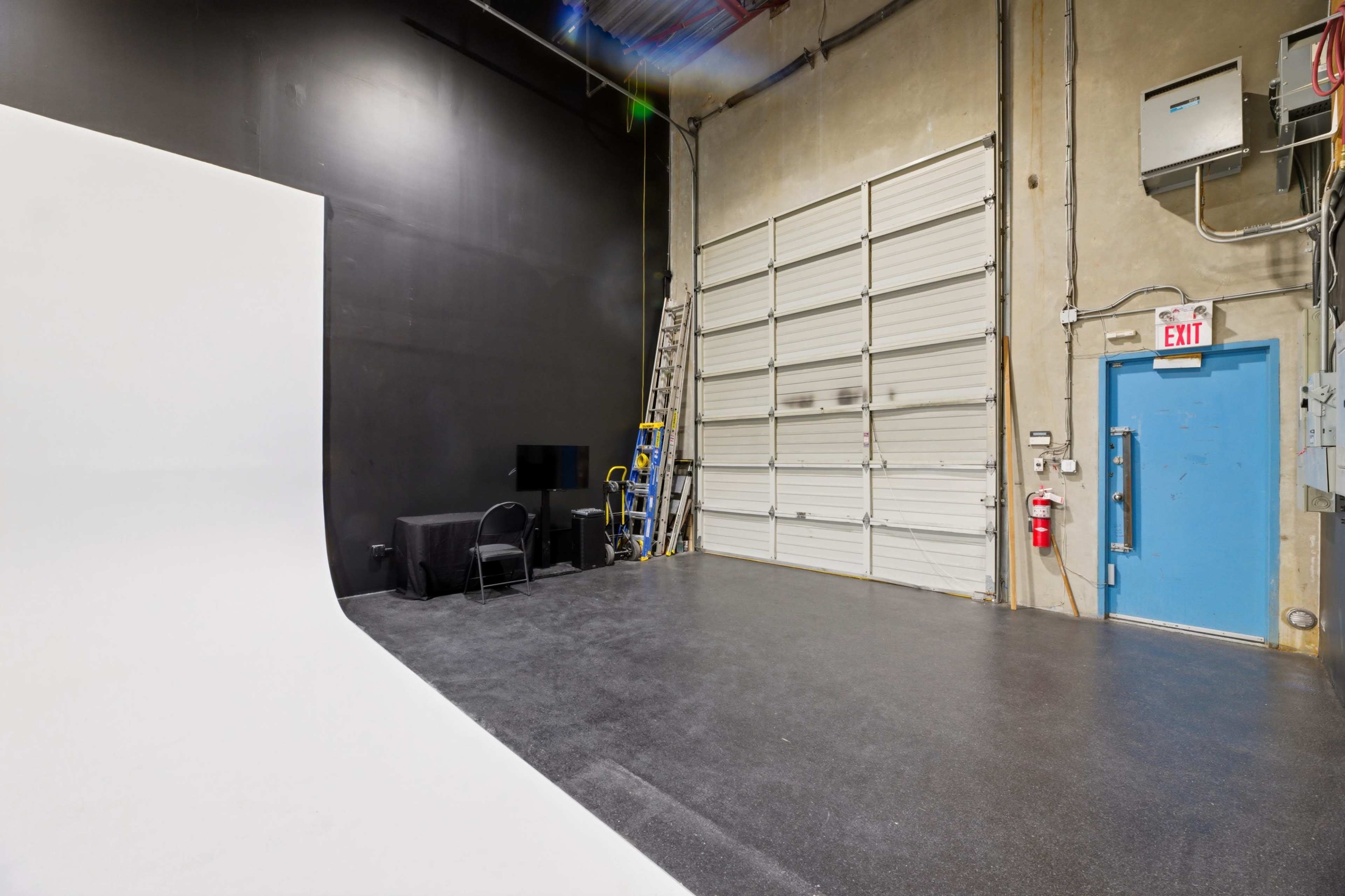 The image shows a photography studio with a white backdrop on one side, a black wall, and a large garage door beside a blue exit door.