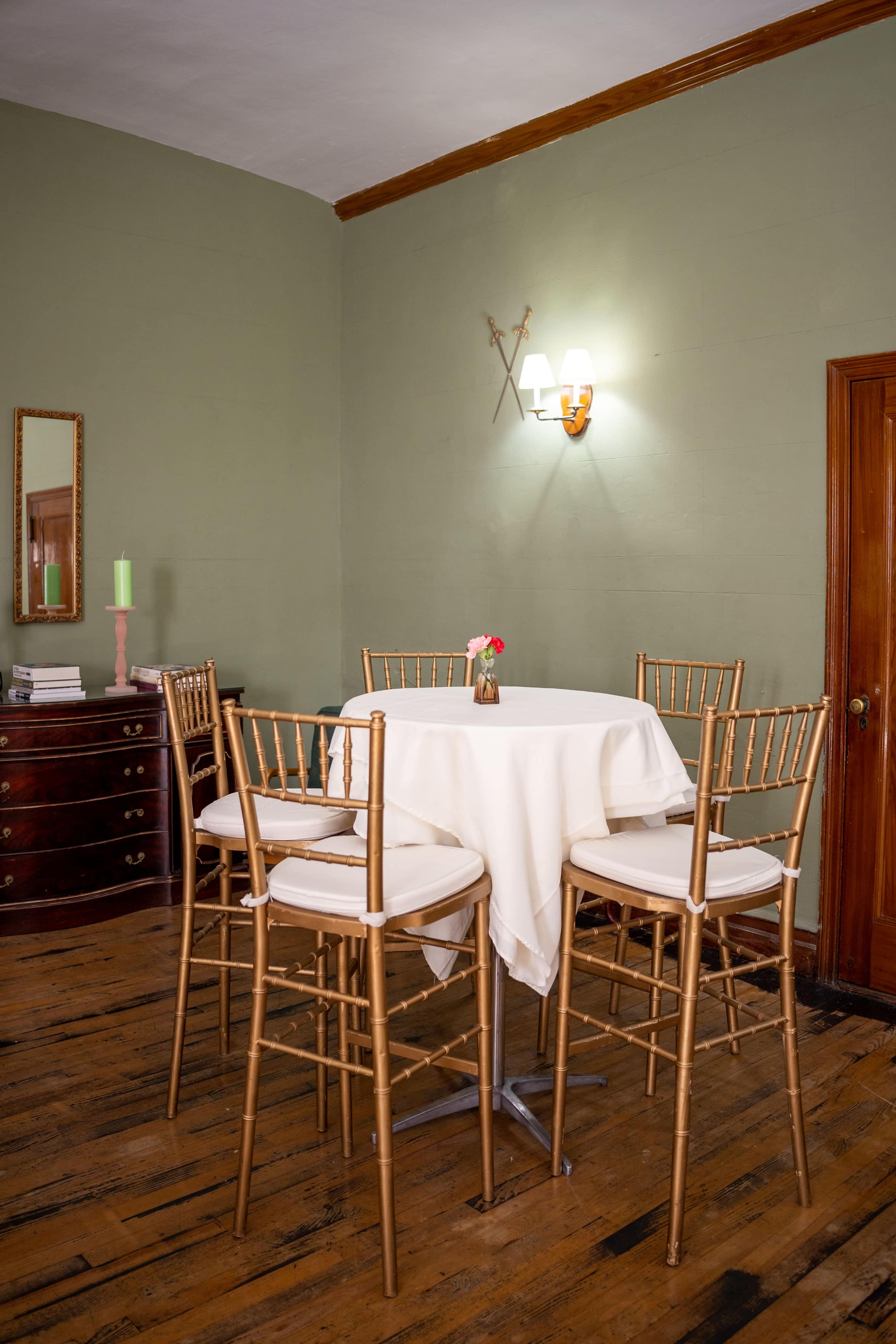 The image shows a small round table covered with a white tablecloth, surrounded by high chairs, in a room with green walls and wooden flooring.