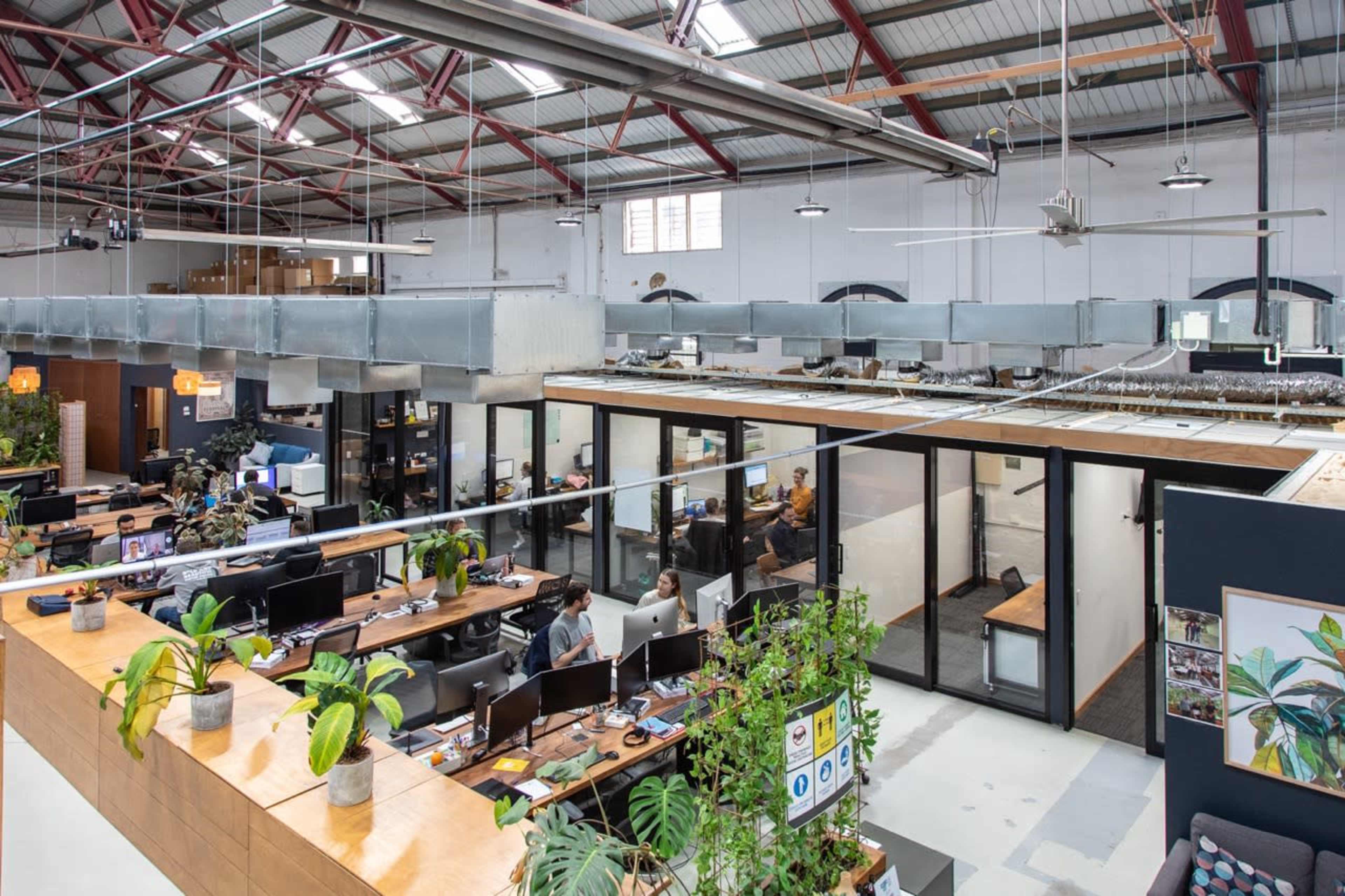 The image shows an open office space with workstations arranged along long wooden tables and glass-enclosed meeting rooms in a large industrial building.