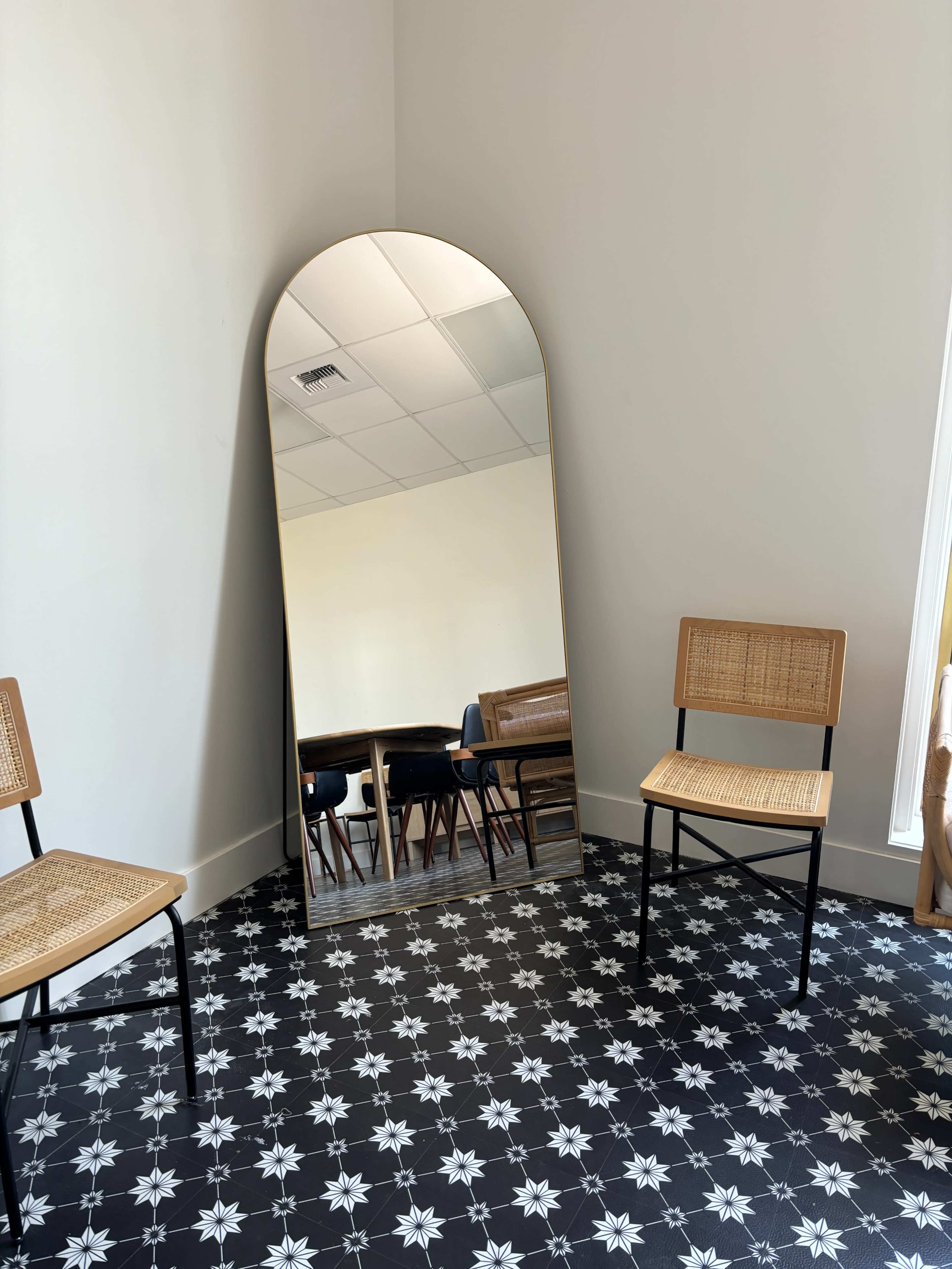 A large arched mirror leans against a wall beside two wooden chairs, set on a patterned black and white floor.