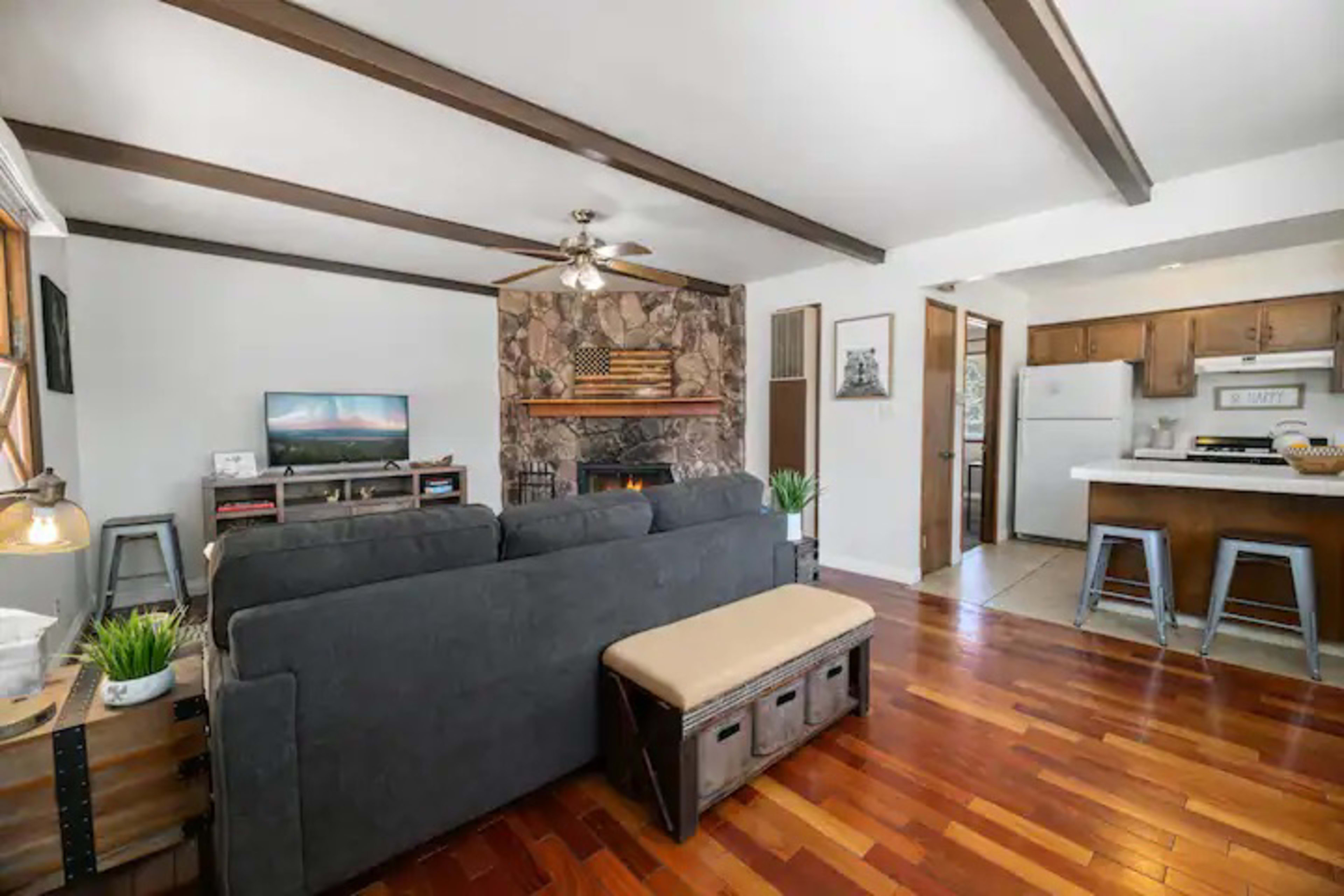 A living room with a stone fireplace, a television on a stand, and a kitchen area visible in the background.