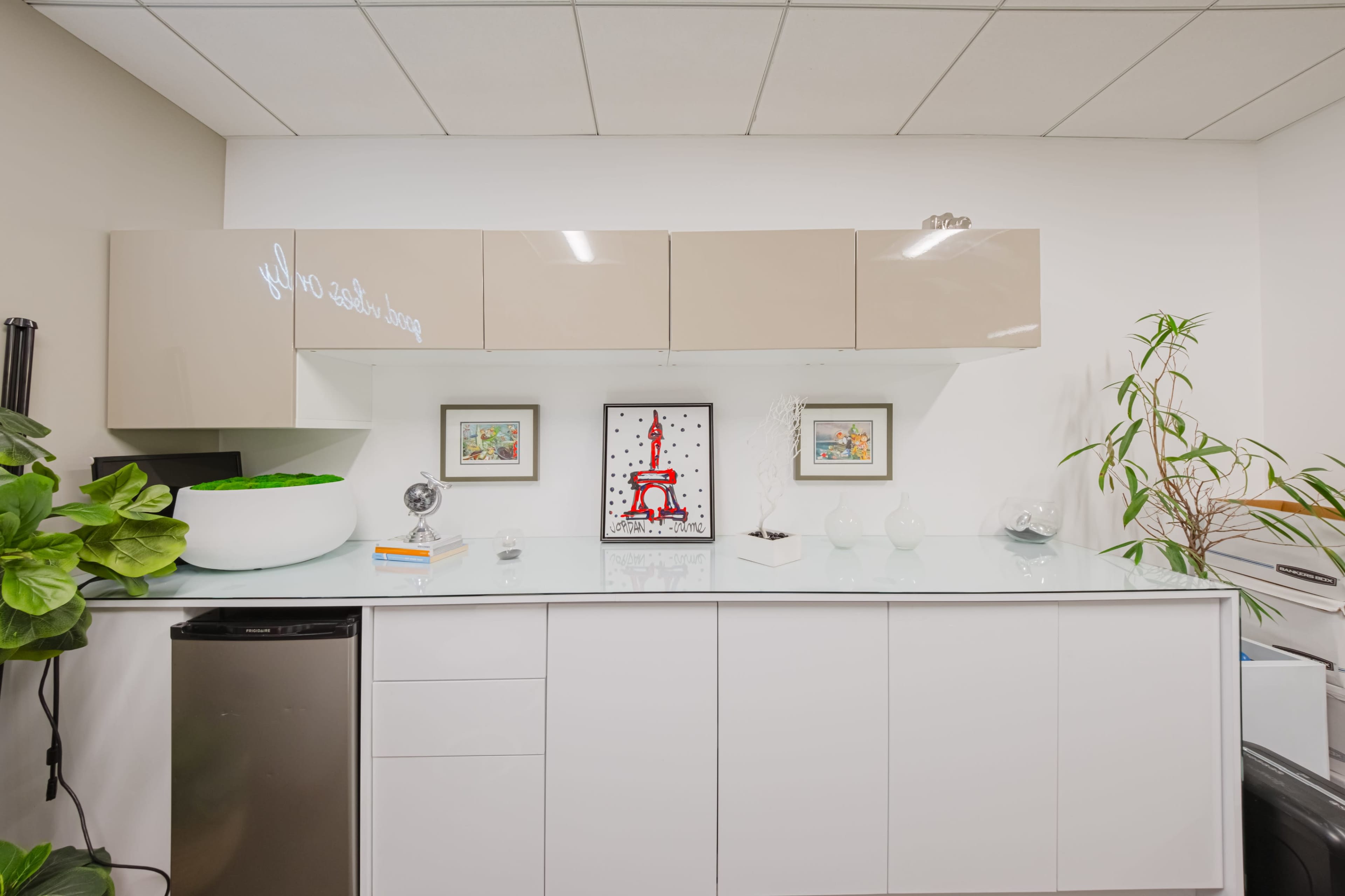 A modern office kitchenette with a white countertop, light-colored cabinets, a small refrigerator, and decorative wall art.