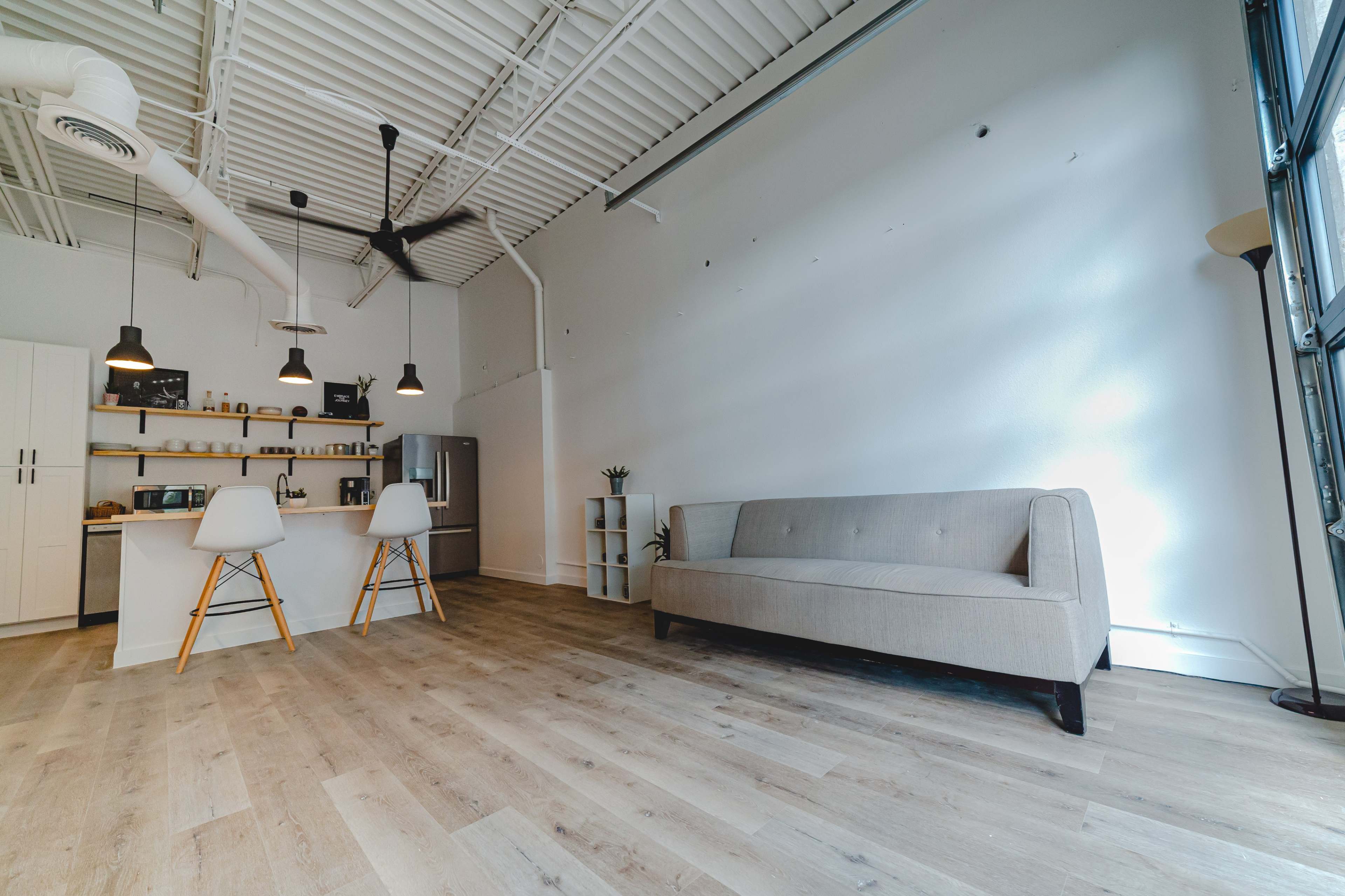 The image shows a minimalistic interior space featuring a light-colored sofa, a kitchen island with stools, and large windows along one wall.