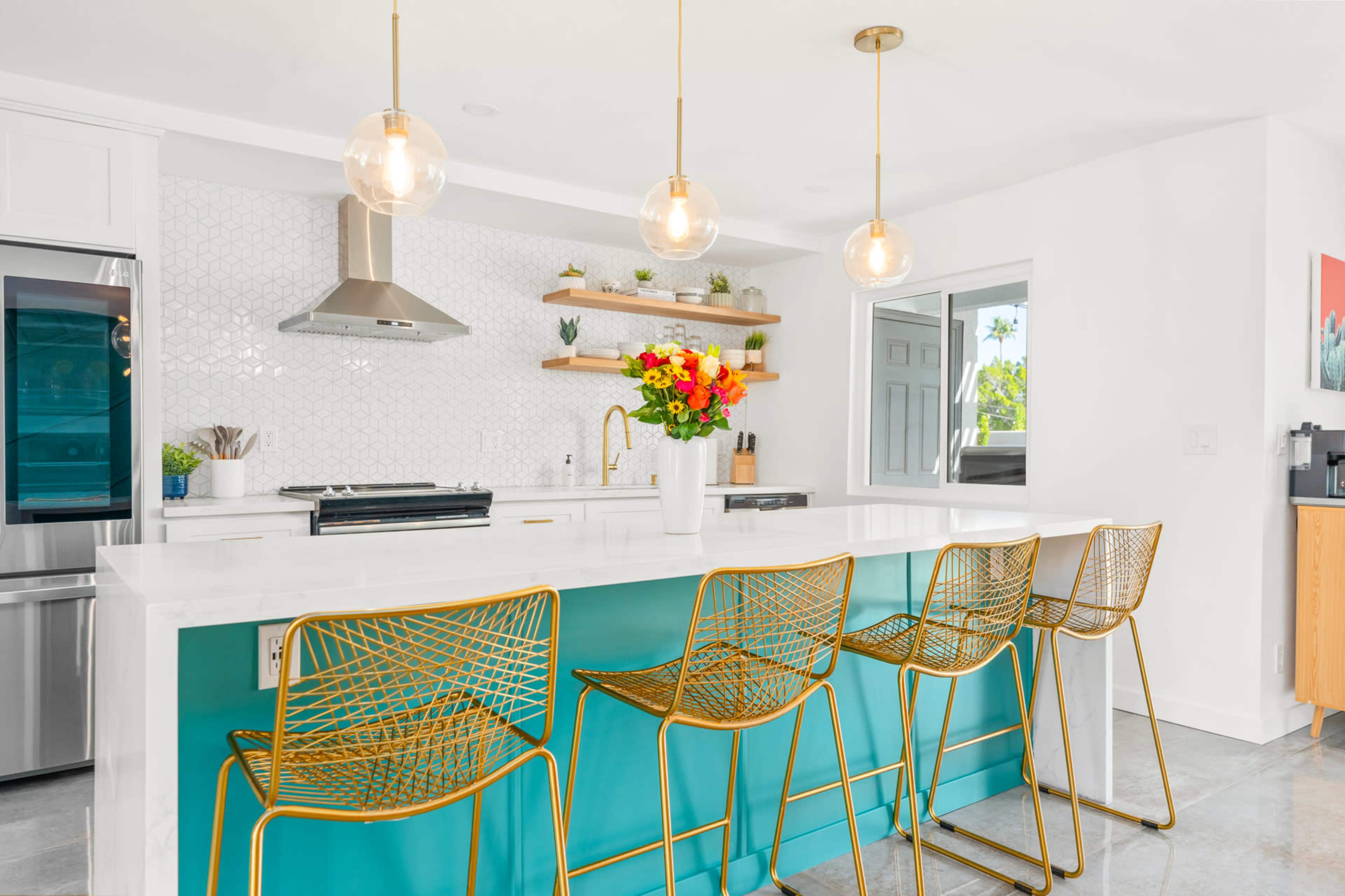 A modern kitchen features a white countertop and teal bar with four gold bar stools, illuminated by three pendant lights.