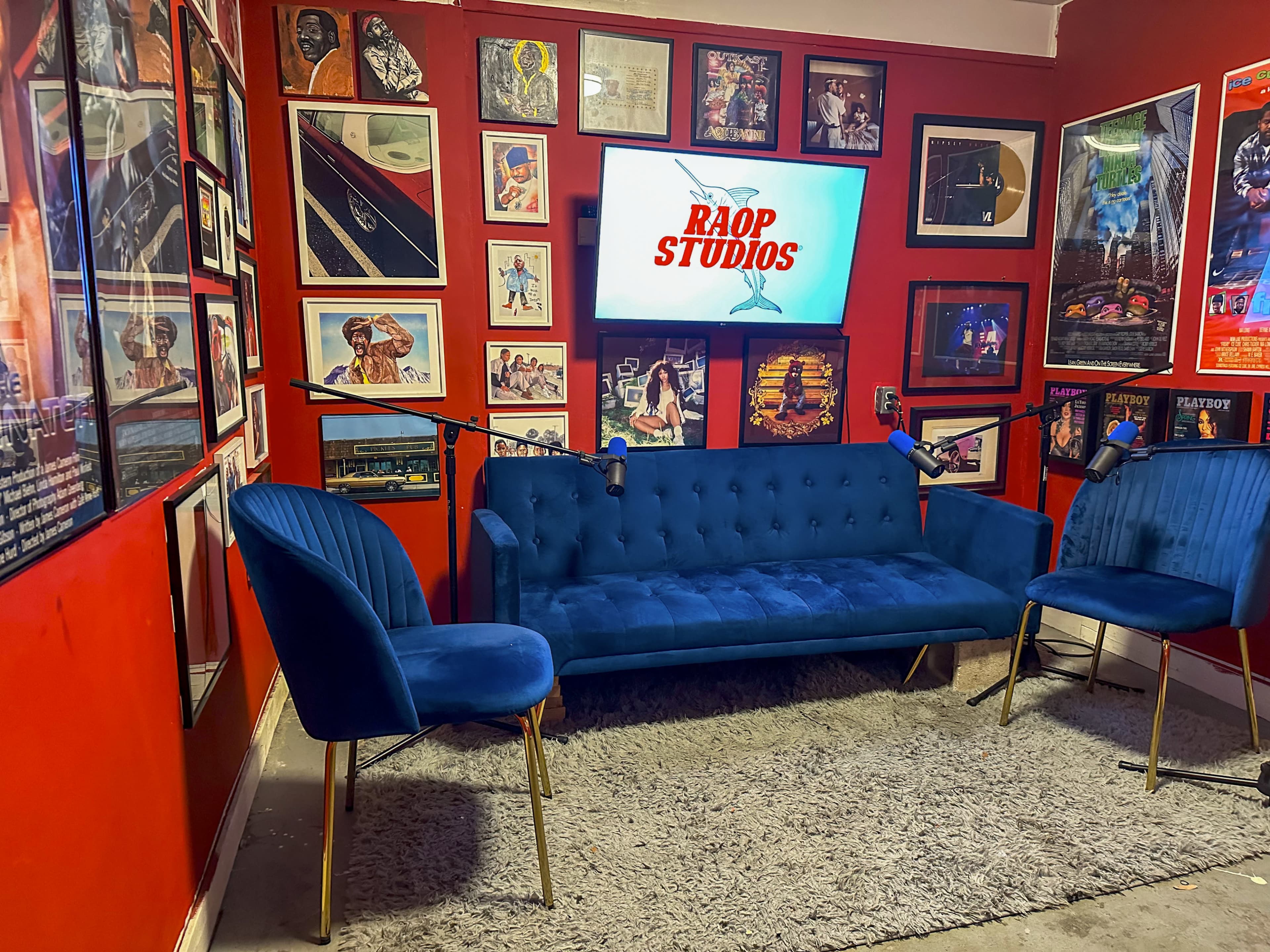 A small recording studio with a red wall adorned with framed posters, a blue sofa, two blue chairs, and a screen displaying "RAOP STUDIOS."