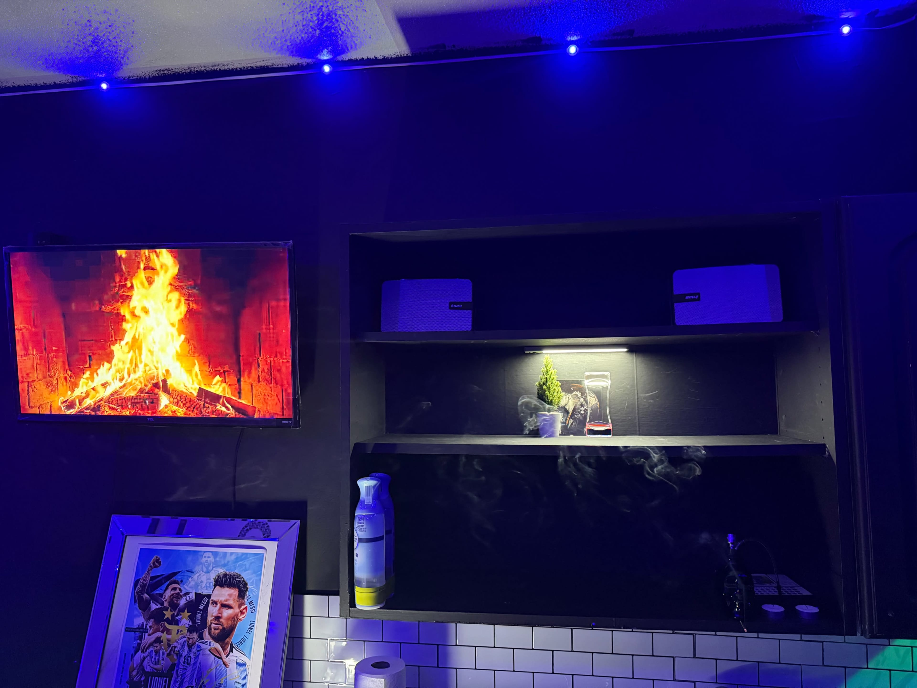 The image shows a cozy interior space featuring a television displaying a fireplace scene, shelves with decorative items, and blue LED strip lighting along the ceiling.