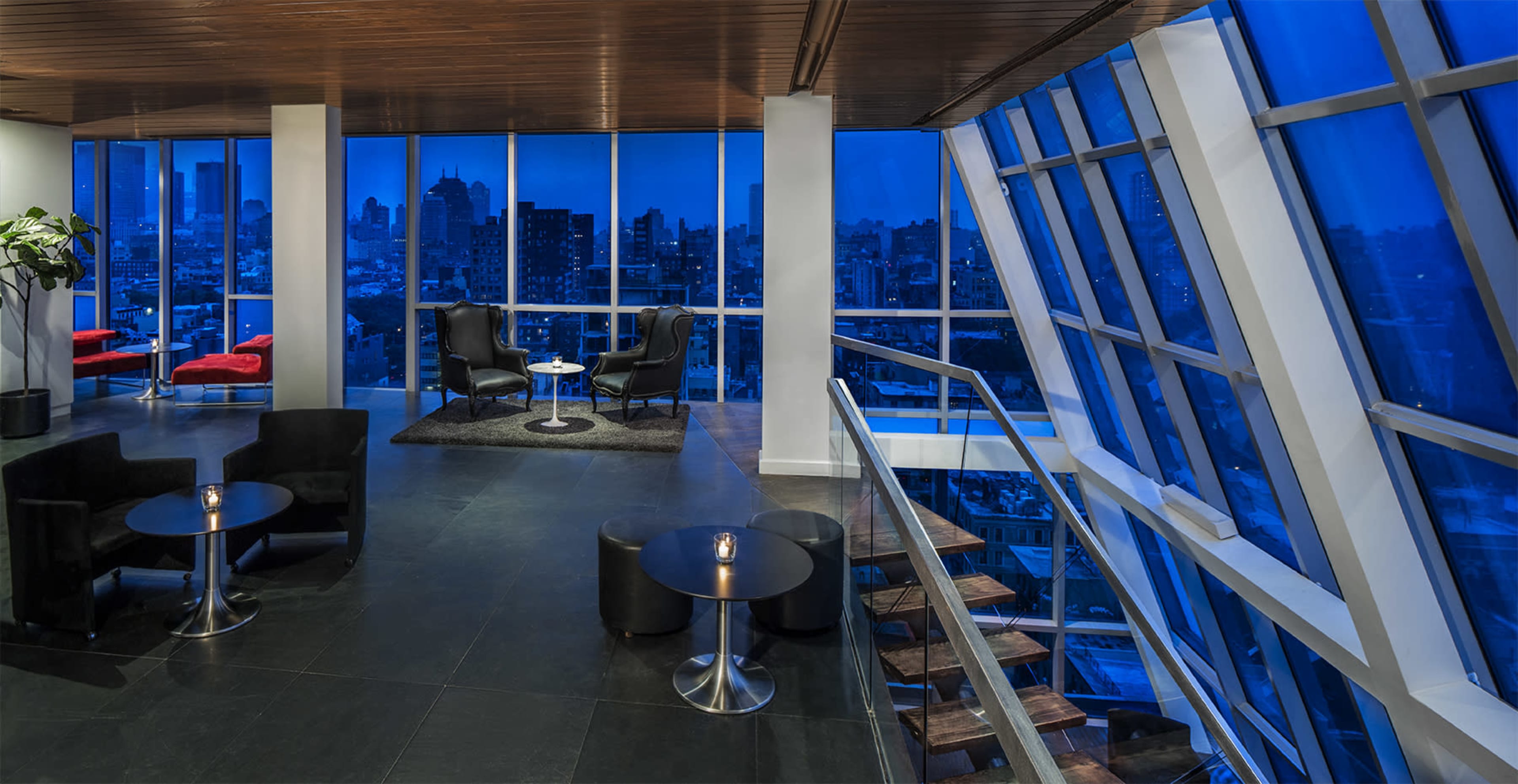 A modern loft-style interior featuring black furniture, a small table with a candle, and large windows offering a view of a city skyline at dusk.