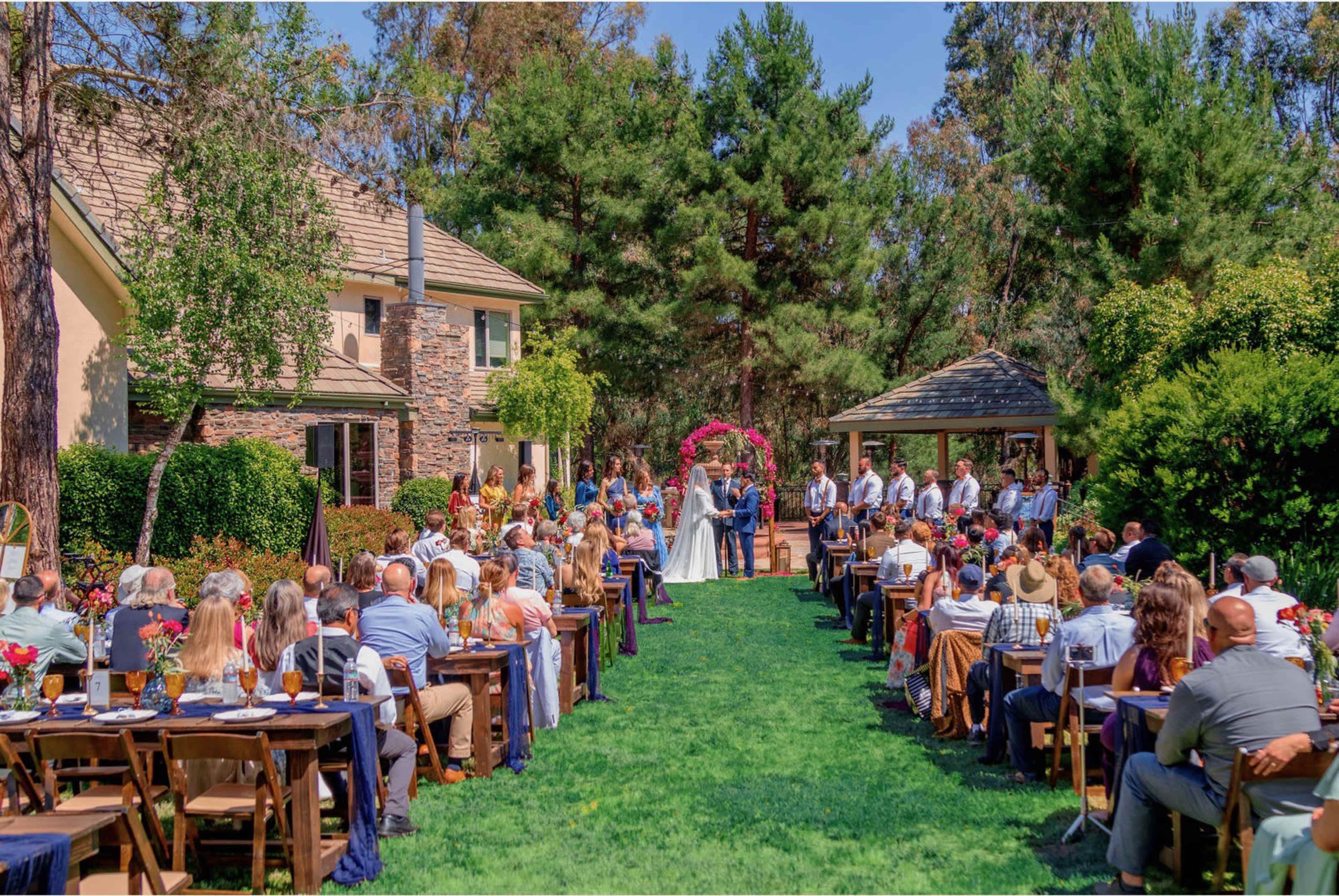 A wedding ceremony takes place outdoors in a garden setting, with guests seated on either side of an aisle leading to the couple at an altar adorned with flowers.