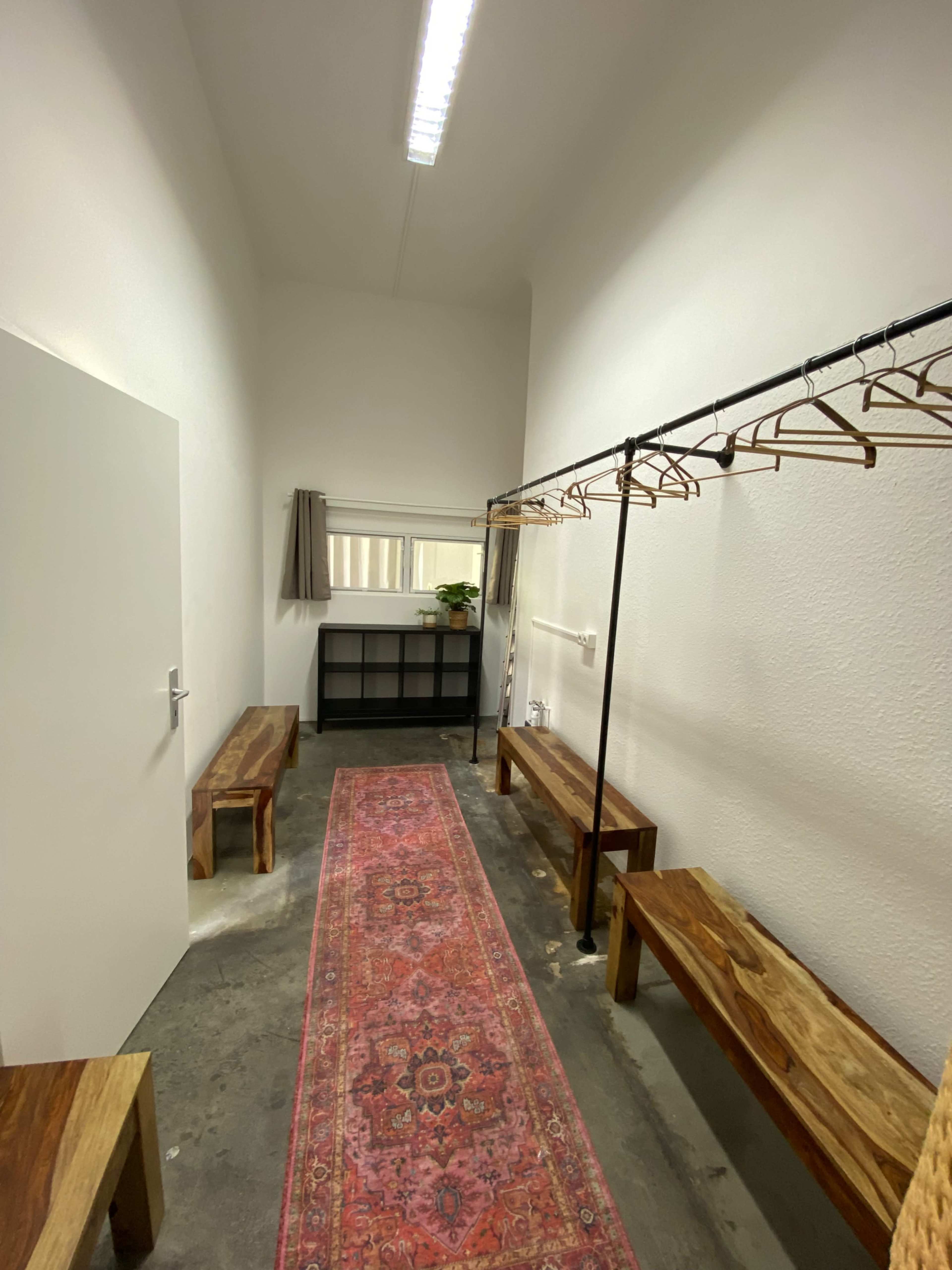 The image shows a narrow room with wooden benches, a hanging rack for clothing, and a long, patterned rug along the floor.