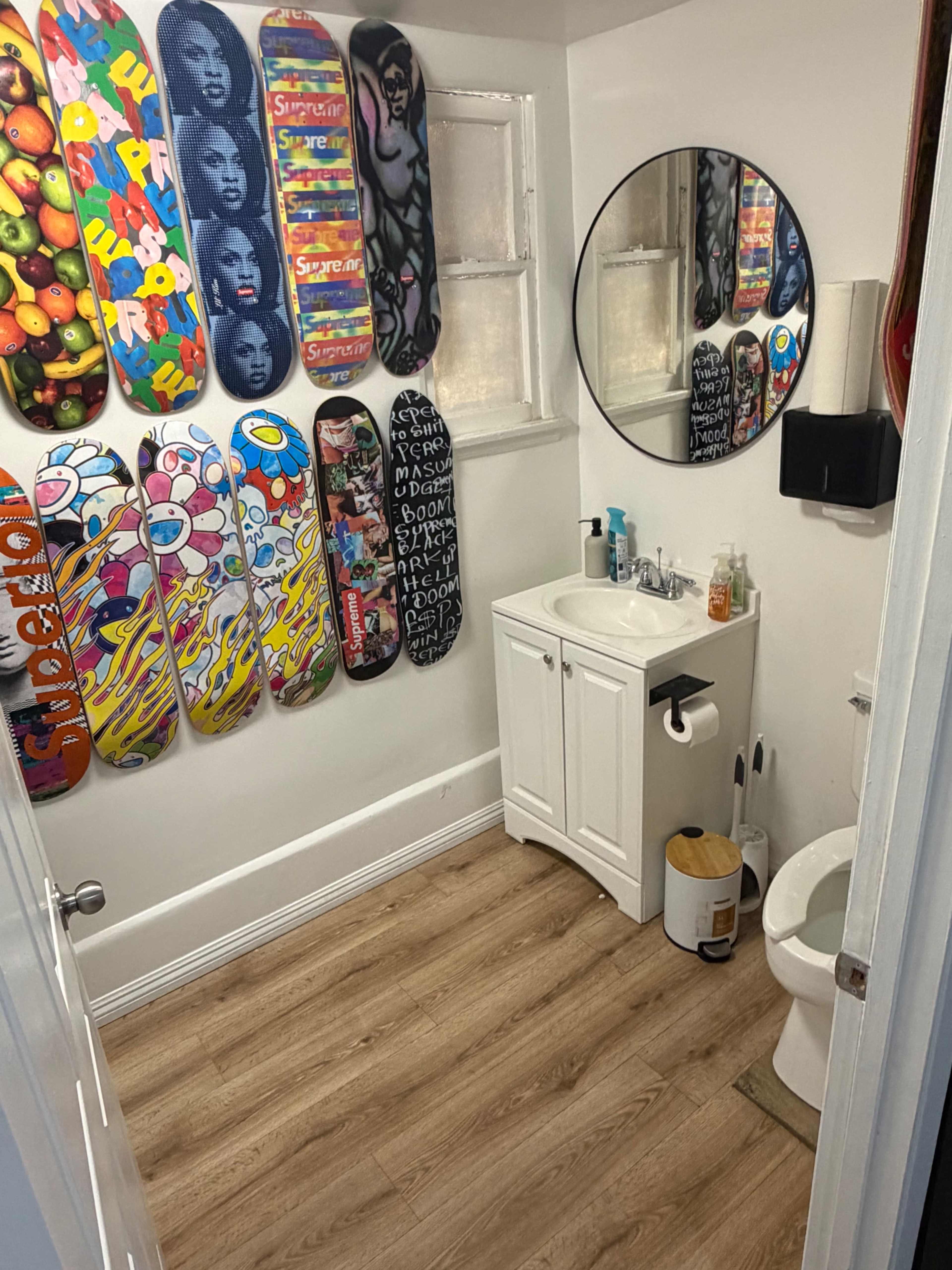 The image shows a small bathroom with a sink, a round mirror, a toilet, and walls adorned with colorful skateboard decks.