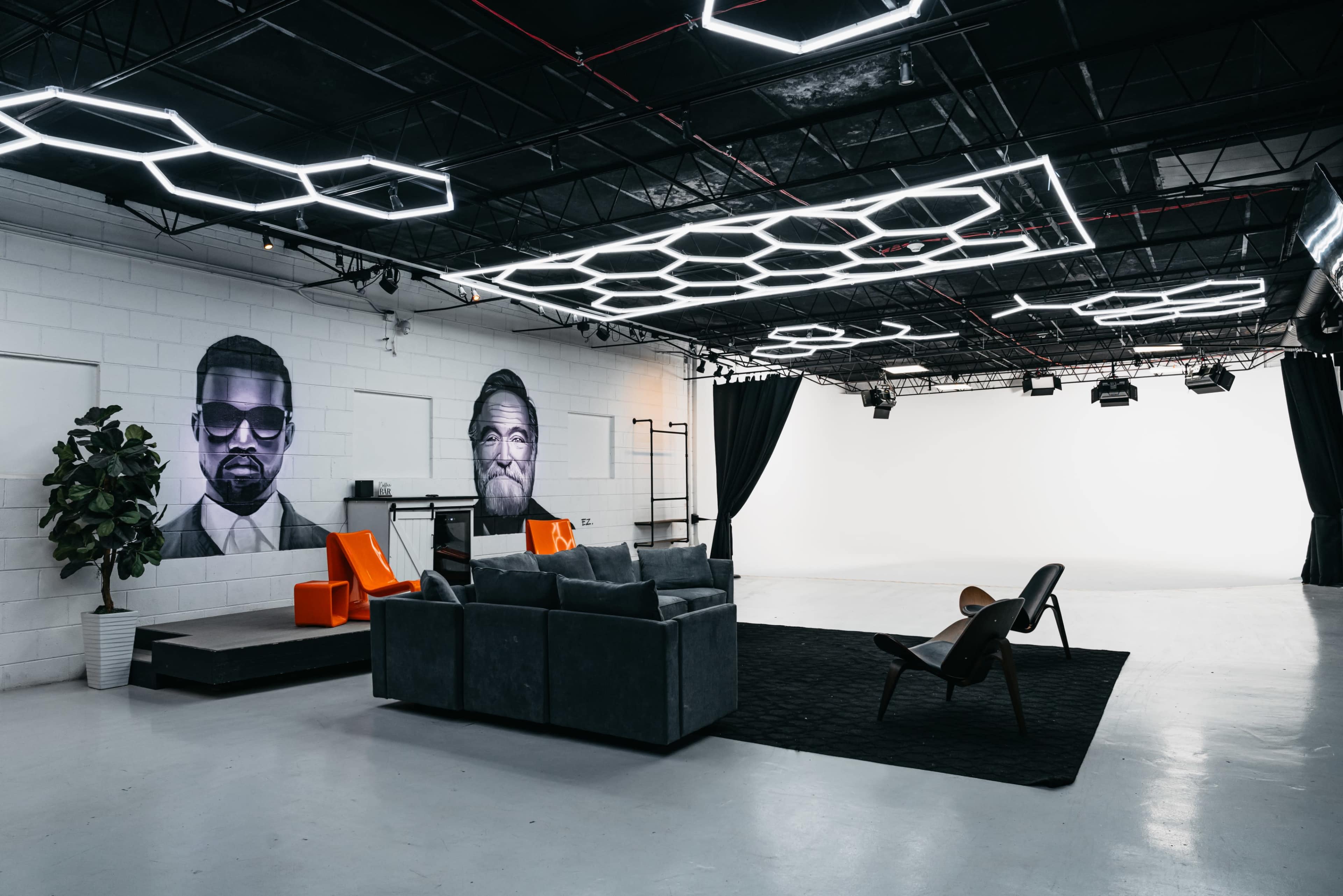 A modern studio space with a minimalist design, showcasing two large portraits on the wall, a seating area with a gray couch and chairs, and illuminated hexagonal light fixtures on the ceiling.