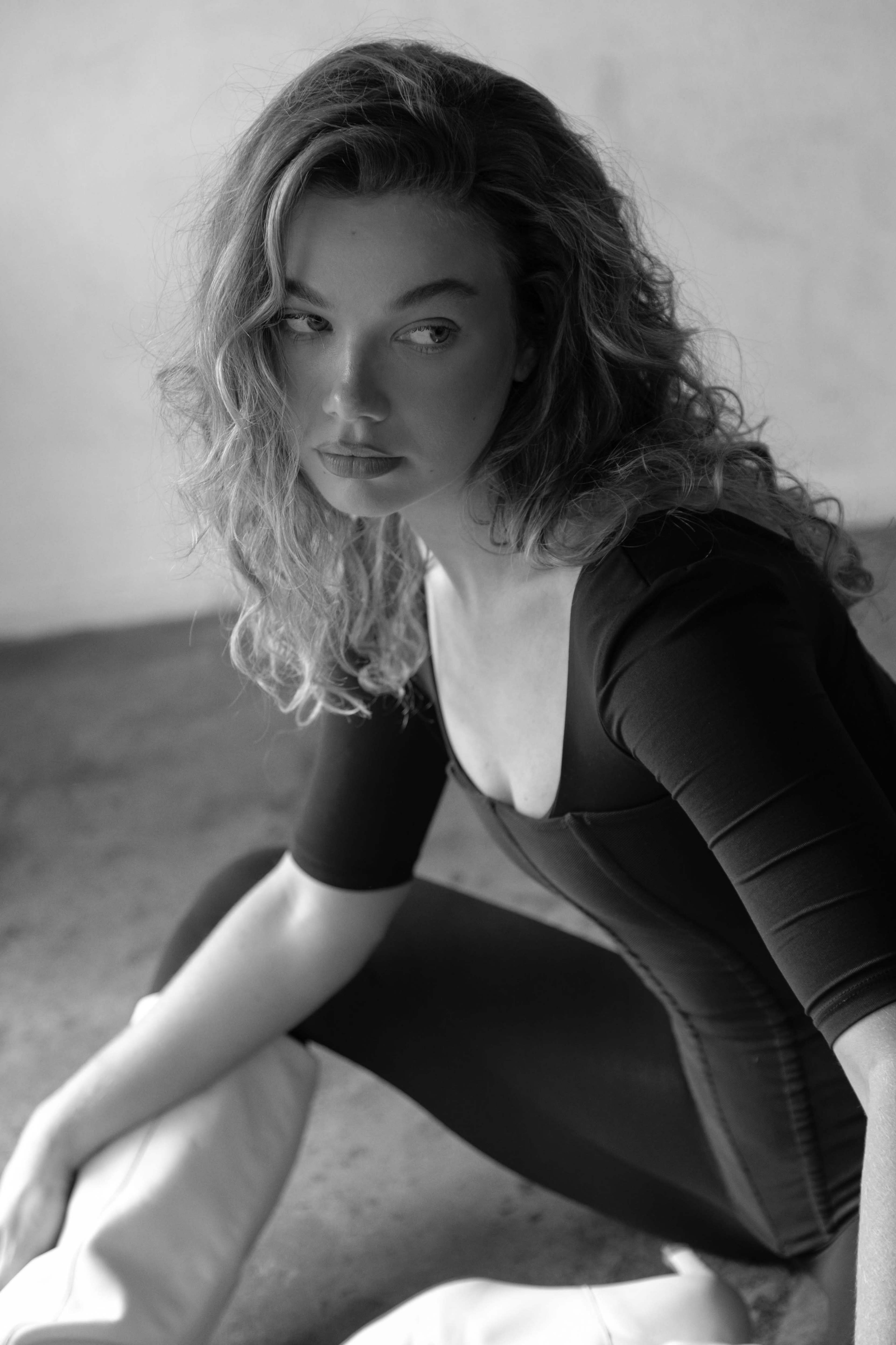 A woman with curly hair sits on a concrete floor, wearing a black fitted top and white pants.