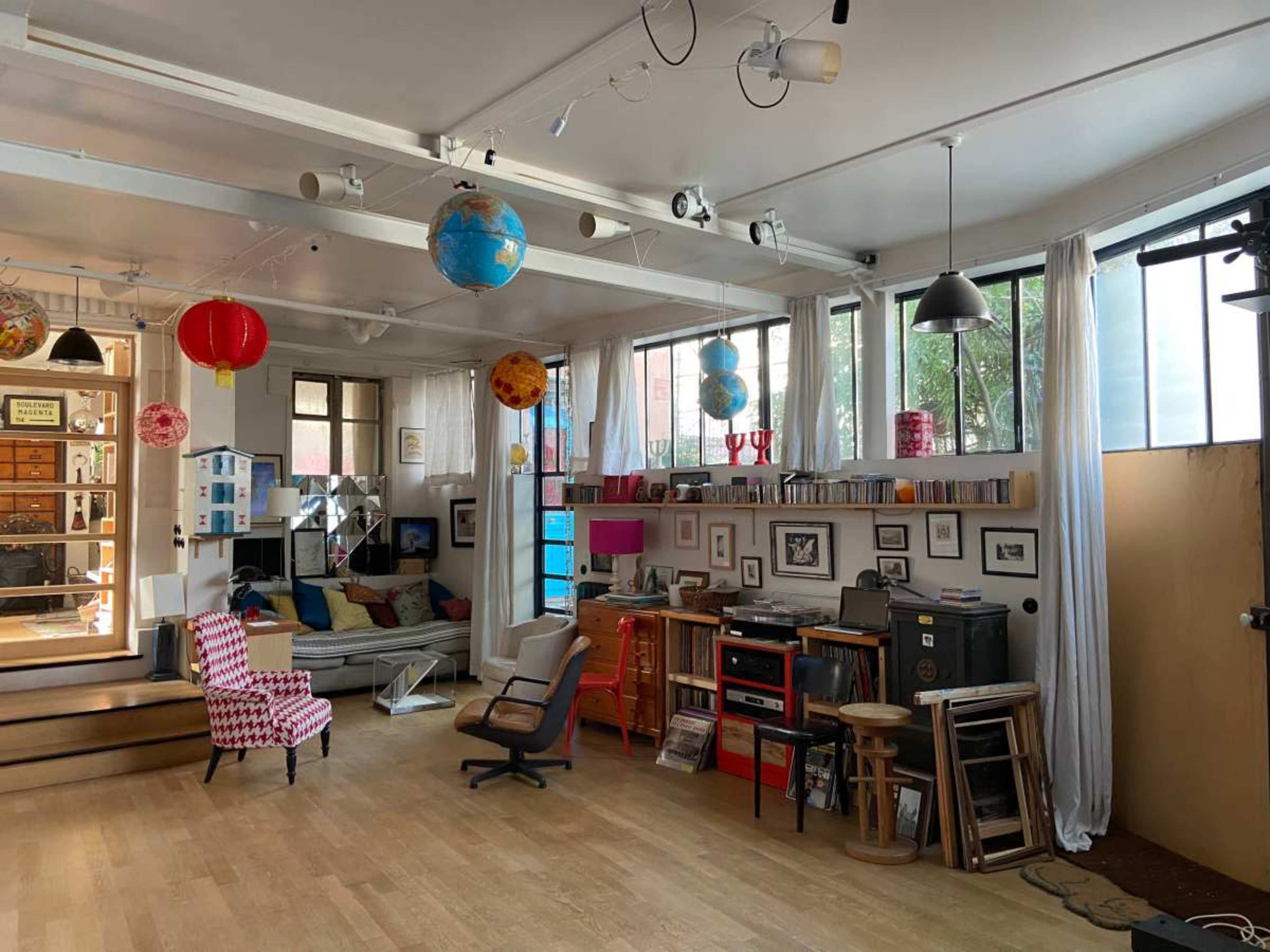 The image shows a spacious and brightly lit living area with large windows, various colorful hanging globes, and a mix of furniture including a striped chair and a wooden stool.