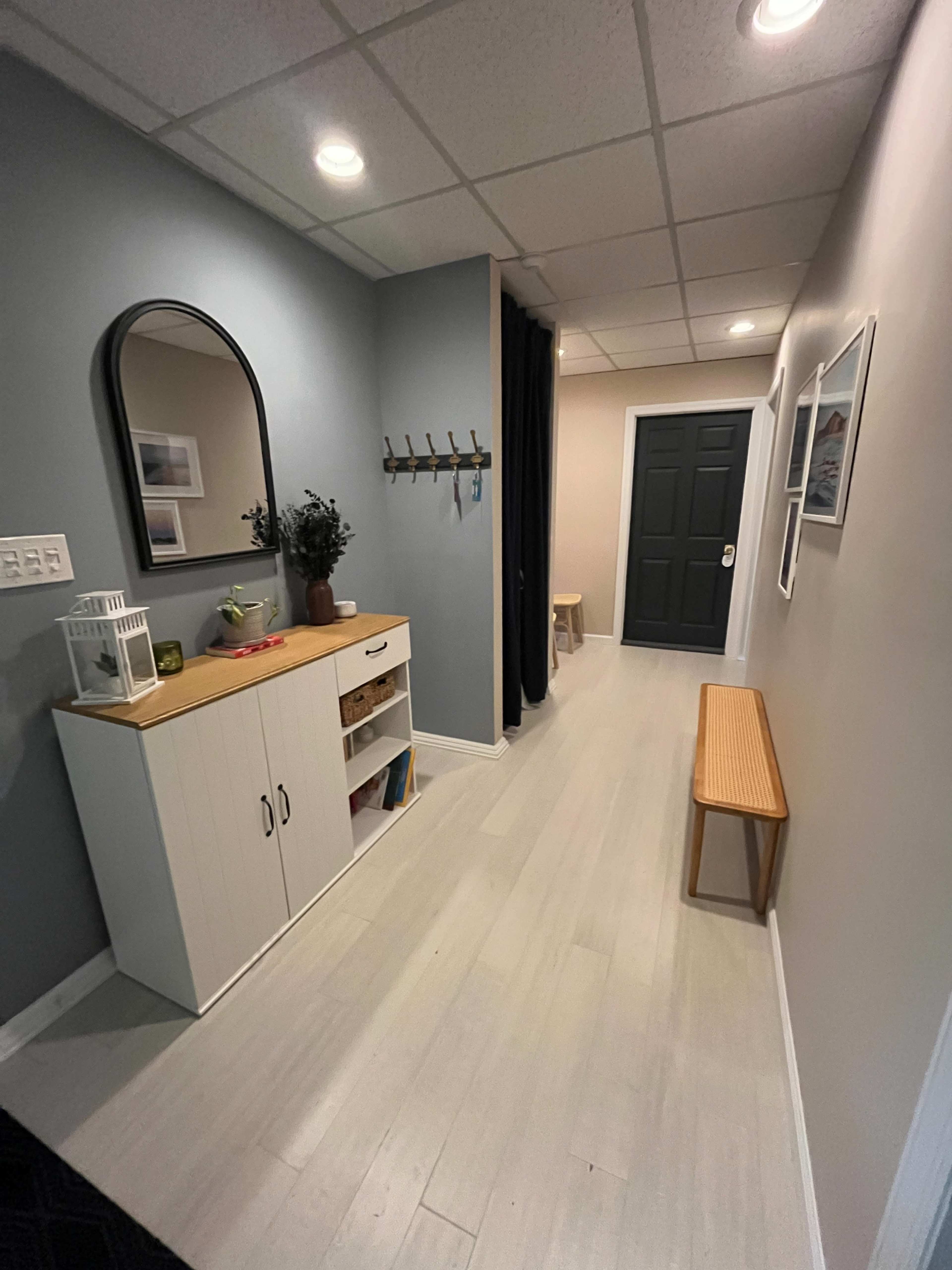 The image shows a narrow hallway featuring a wall-mounted mirror, a cabinet with storage, a coat rack, and a bench along with minimal decorative elements.