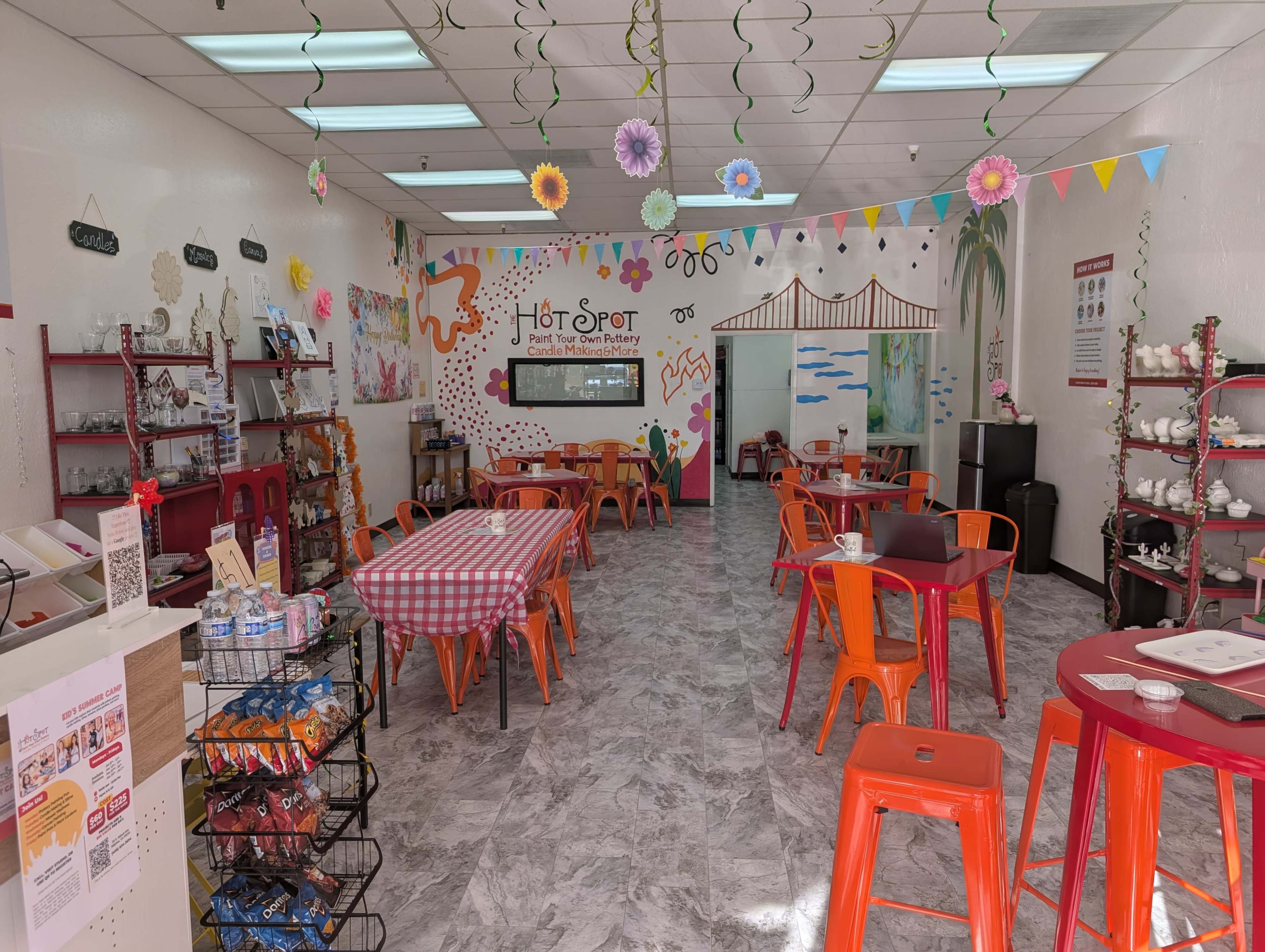 The image shows a brightly decorated café with orange chairs and tables, adorned with colorful paper decorations and a mural on the wall.