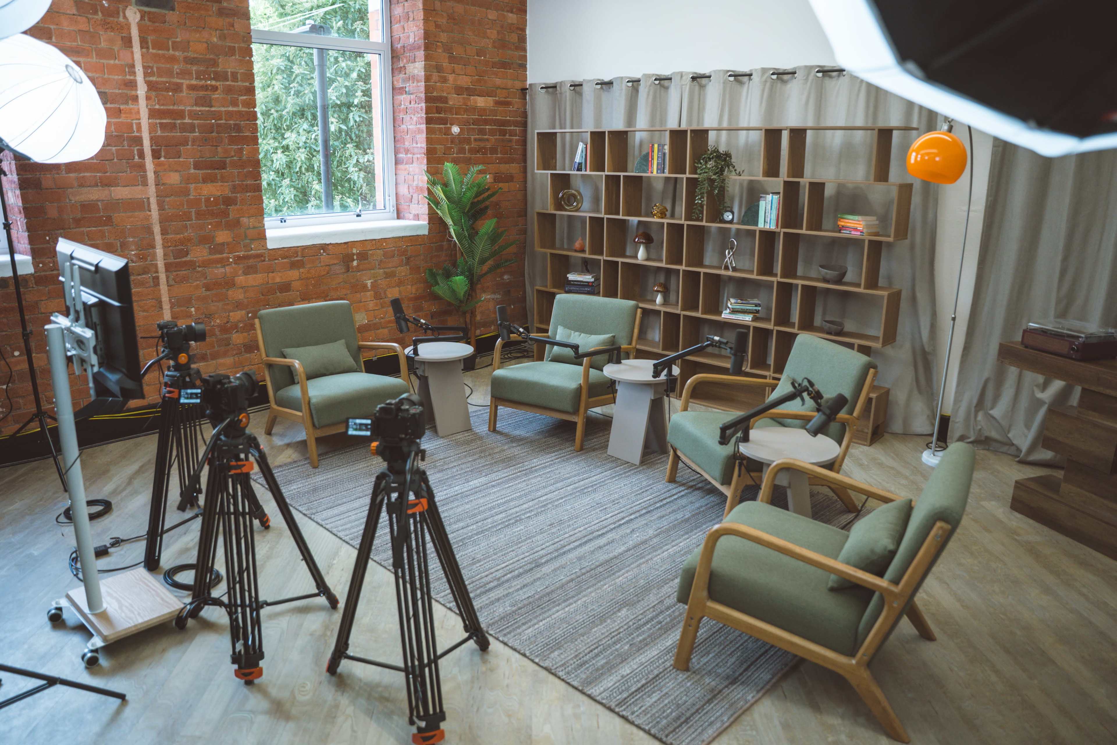 The image shows a modern, well-lit interior space featuring green upholstered chairs arranged around a central table, with several tripods and cameras positioned in front of a bookshelves and window.
