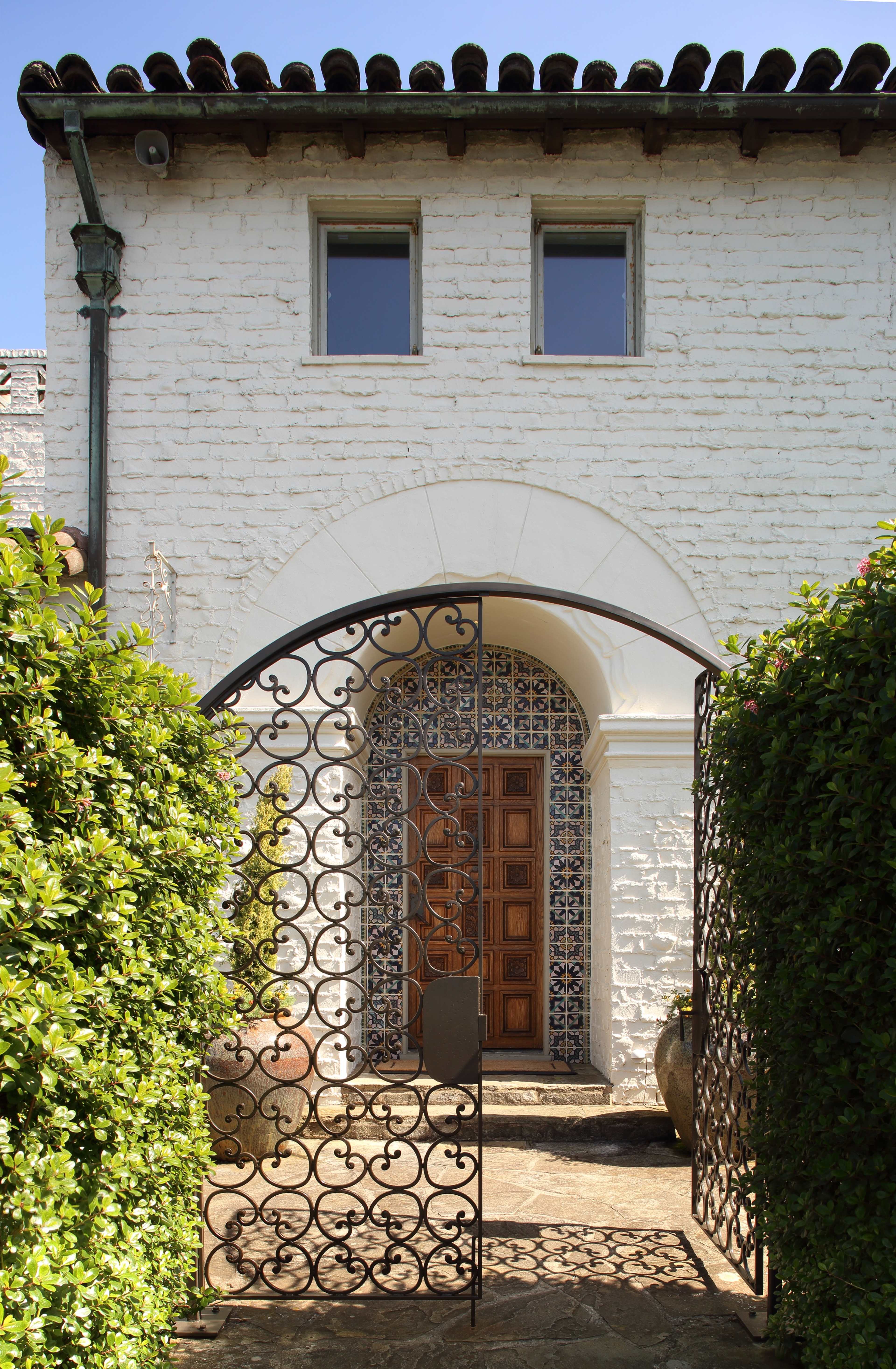 An ornate wrought-iron gate leading to a decorative entrance with tiled walls and a wooden door, framed by green hedges and a white brick facade.