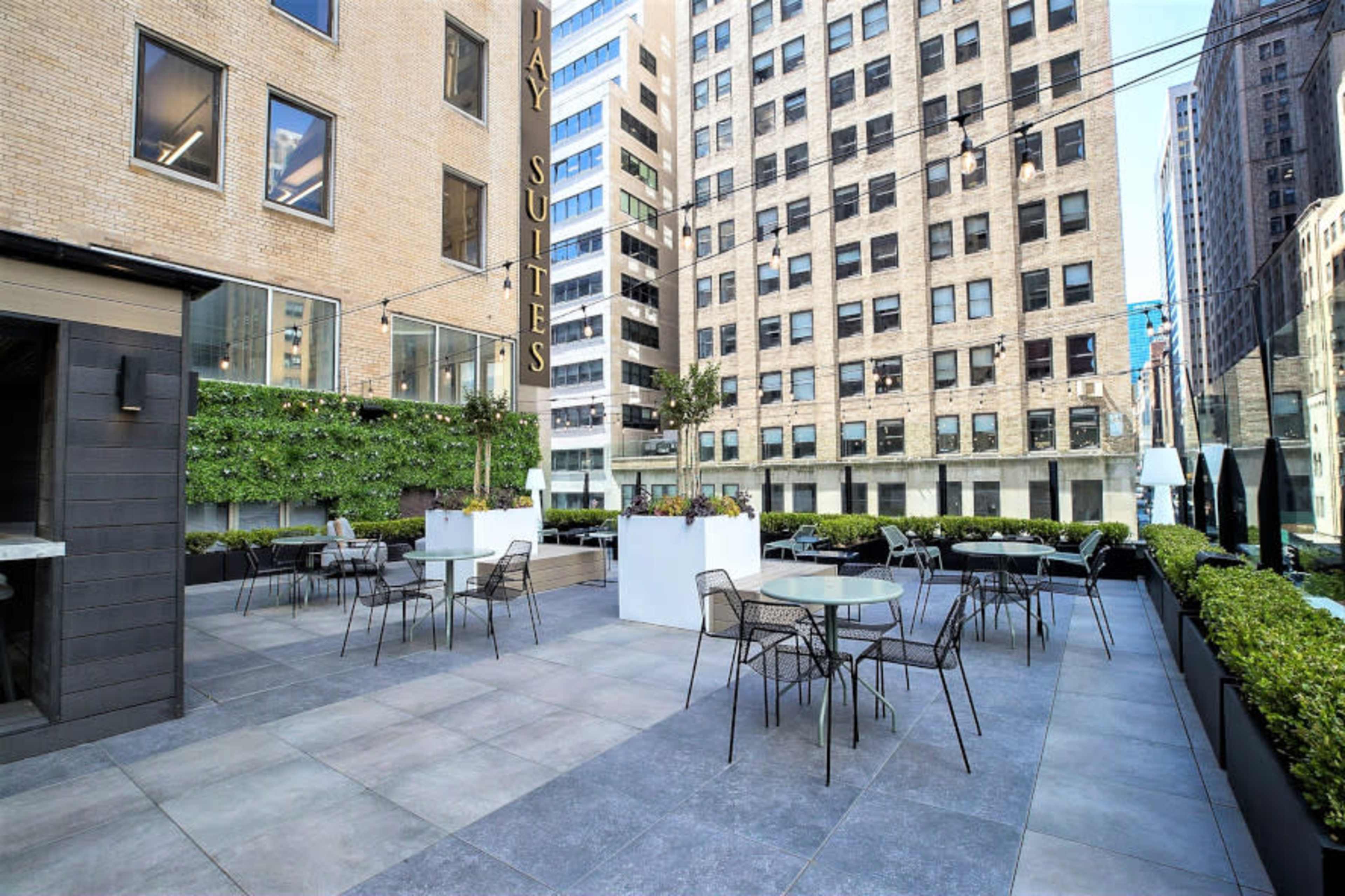 The image shows a modern outdoor terrace area with tables and chairs surrounded by greenery and tall buildings.