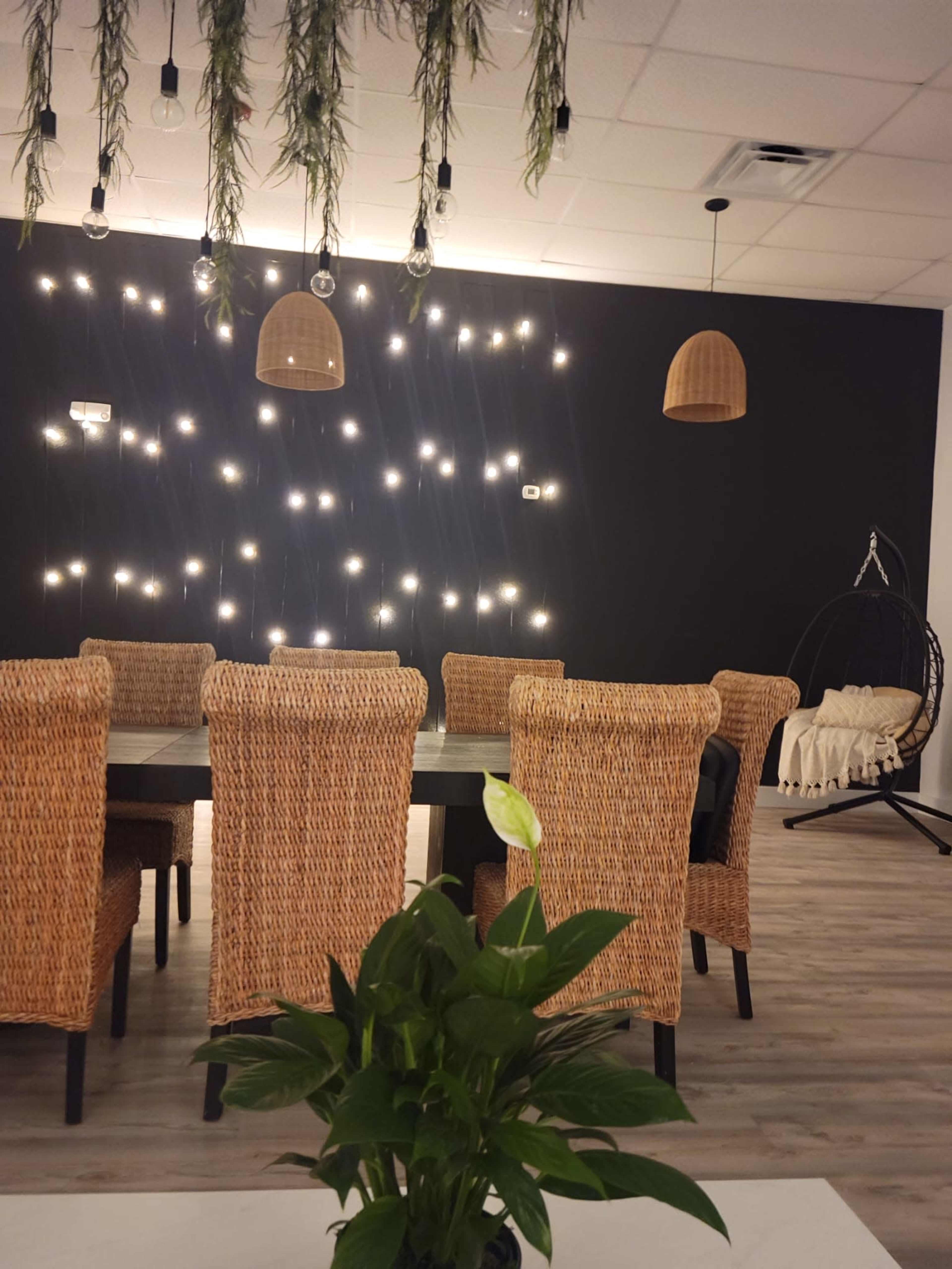 A dining area features wicker chairs around a dark table, with hanging lights and a plant in the foreground.