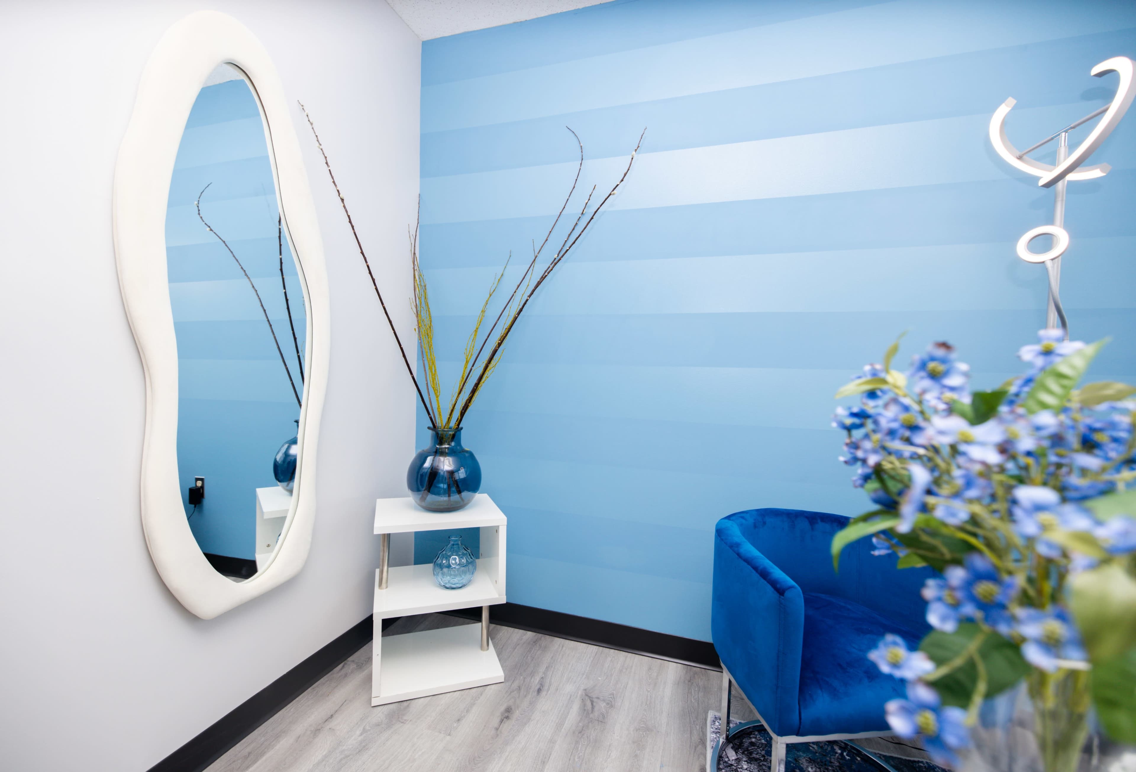 A small room with blue striped walls, a decorative mirror, a shelf holding a vase with branches, and a blue armchair beside a floral arrangement.