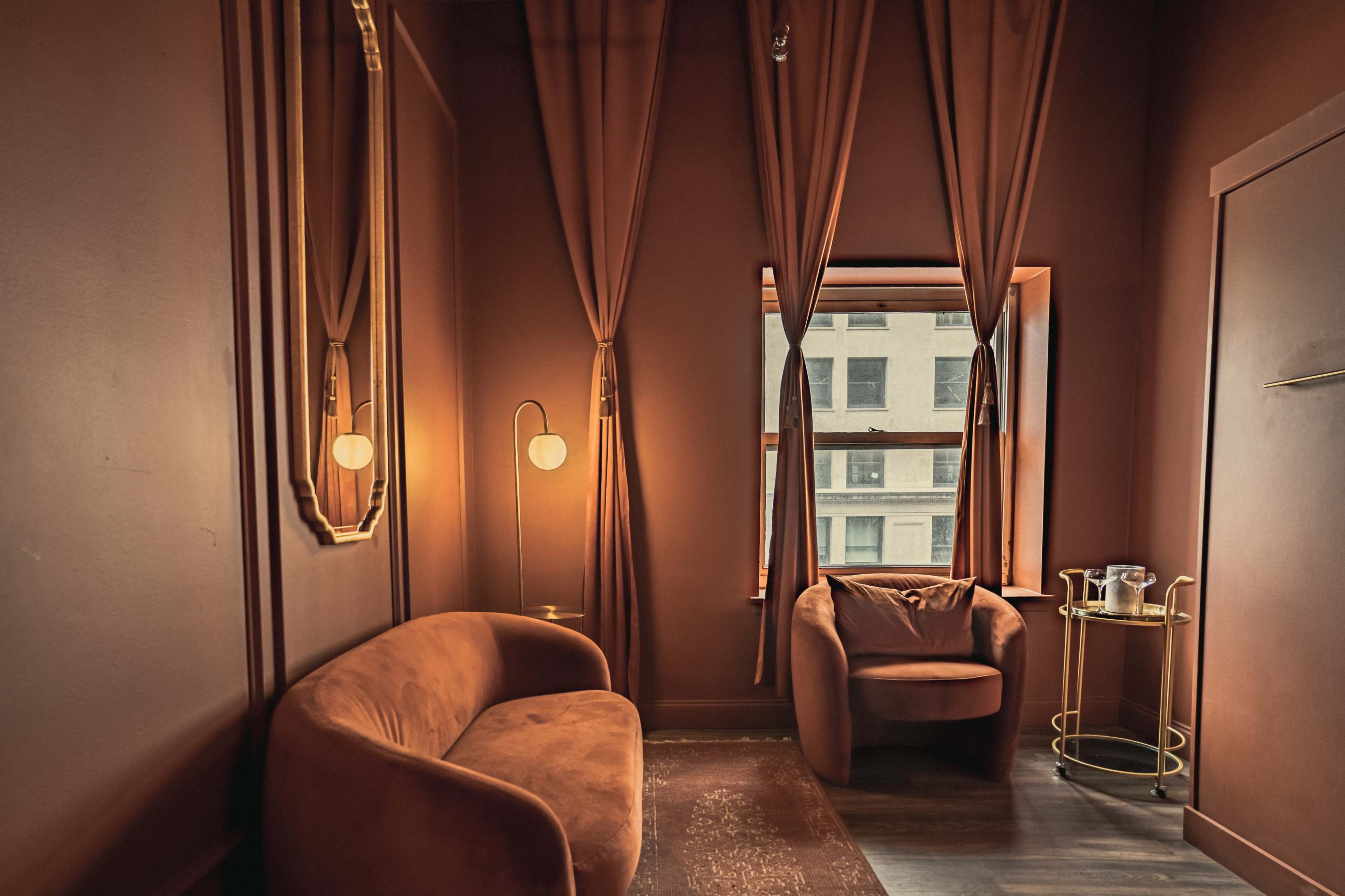 A small, brown-toned room featuring a curved sofa, an armchair, floor lamps, and a window with brown curtains.