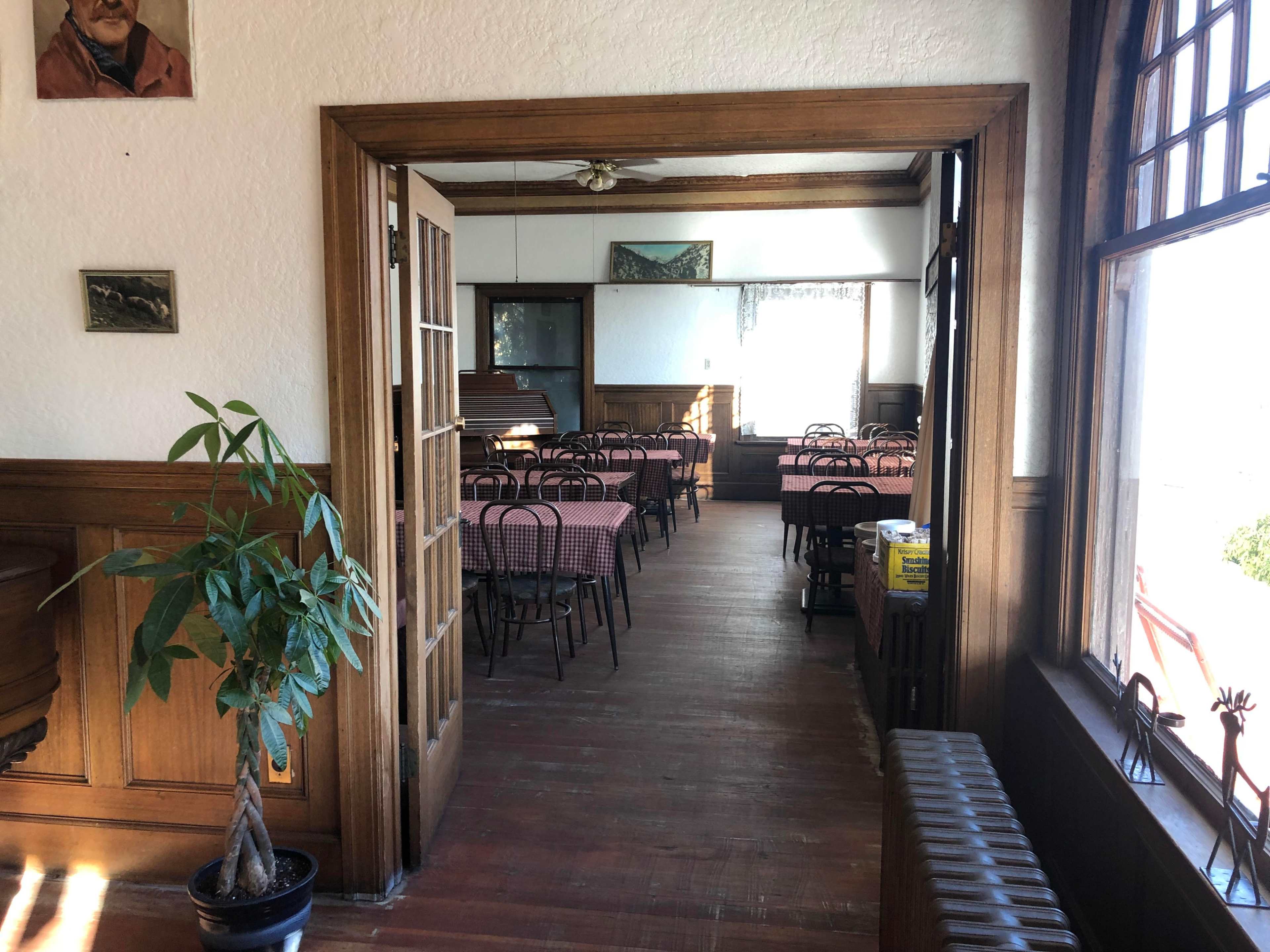 The image shows an empty dining area with checkered tablecloths and wooden furniture, featuring large windows and wooden paneling.