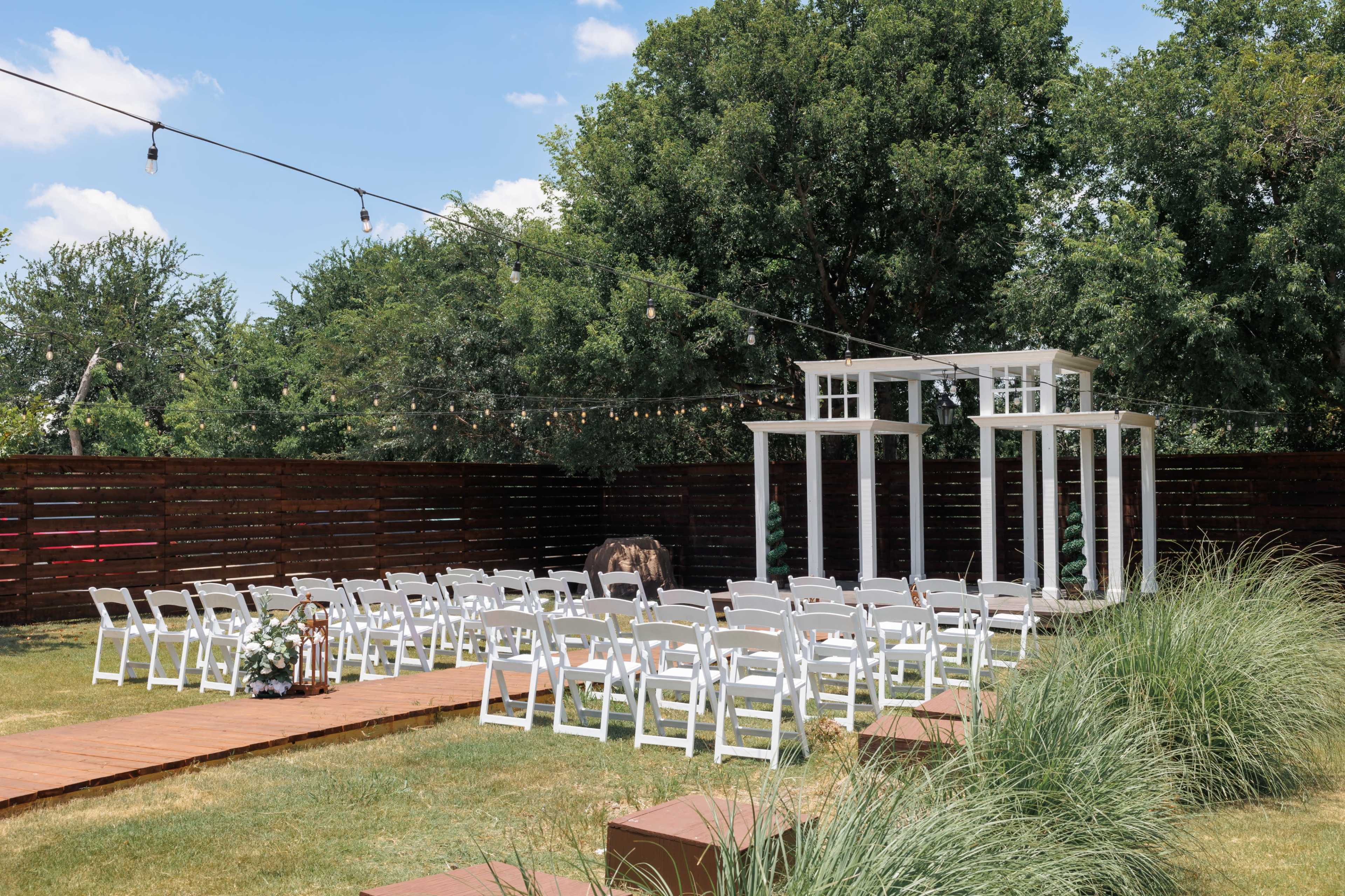An outdoor wedding venue features white chairs arranged in rows facing a decorative arch under string lights, with lush greenery in the background.