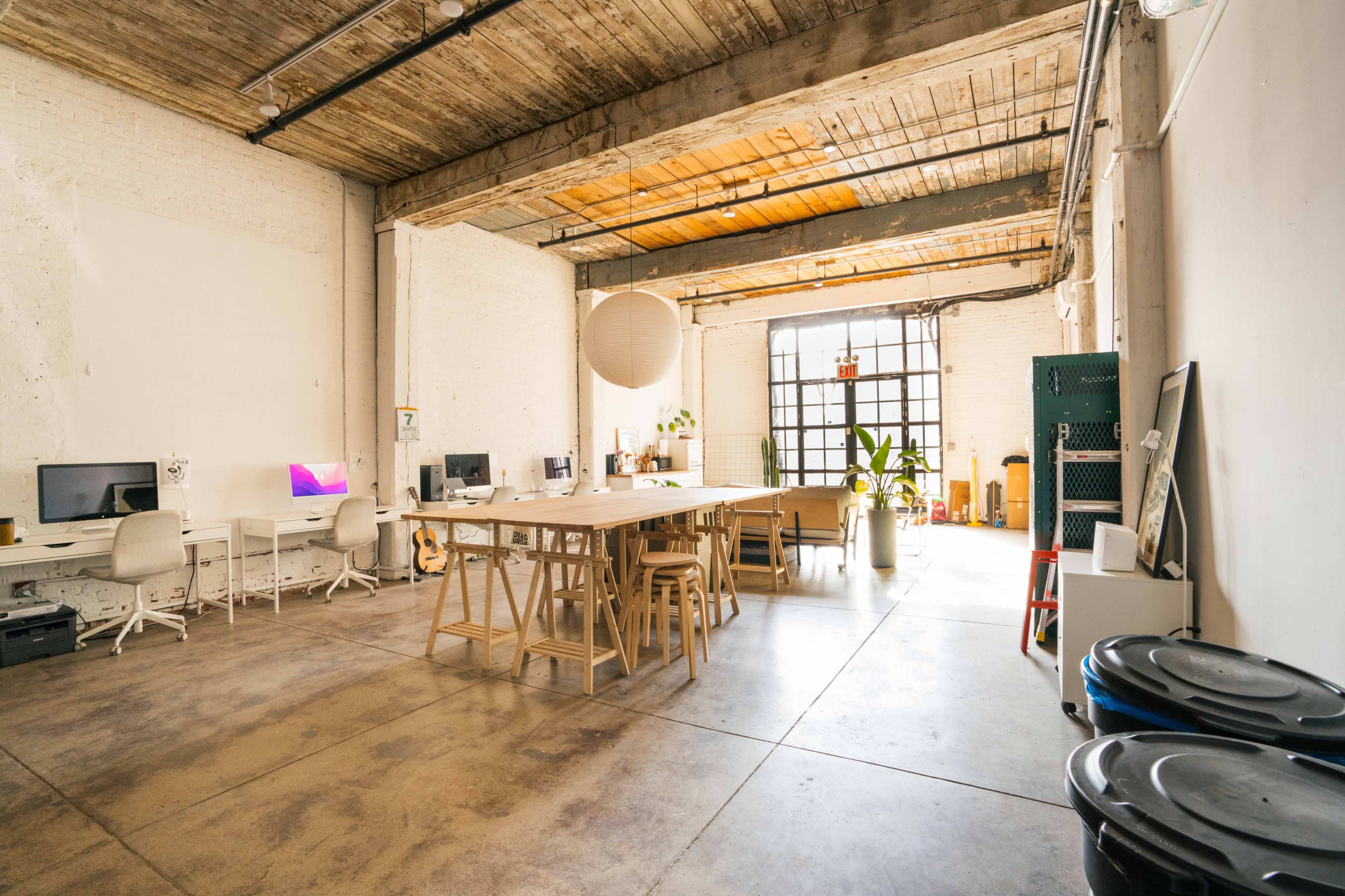 A spacious, well-lit office area with a long wooden table, several workstations, and large windows allowing natural light in.