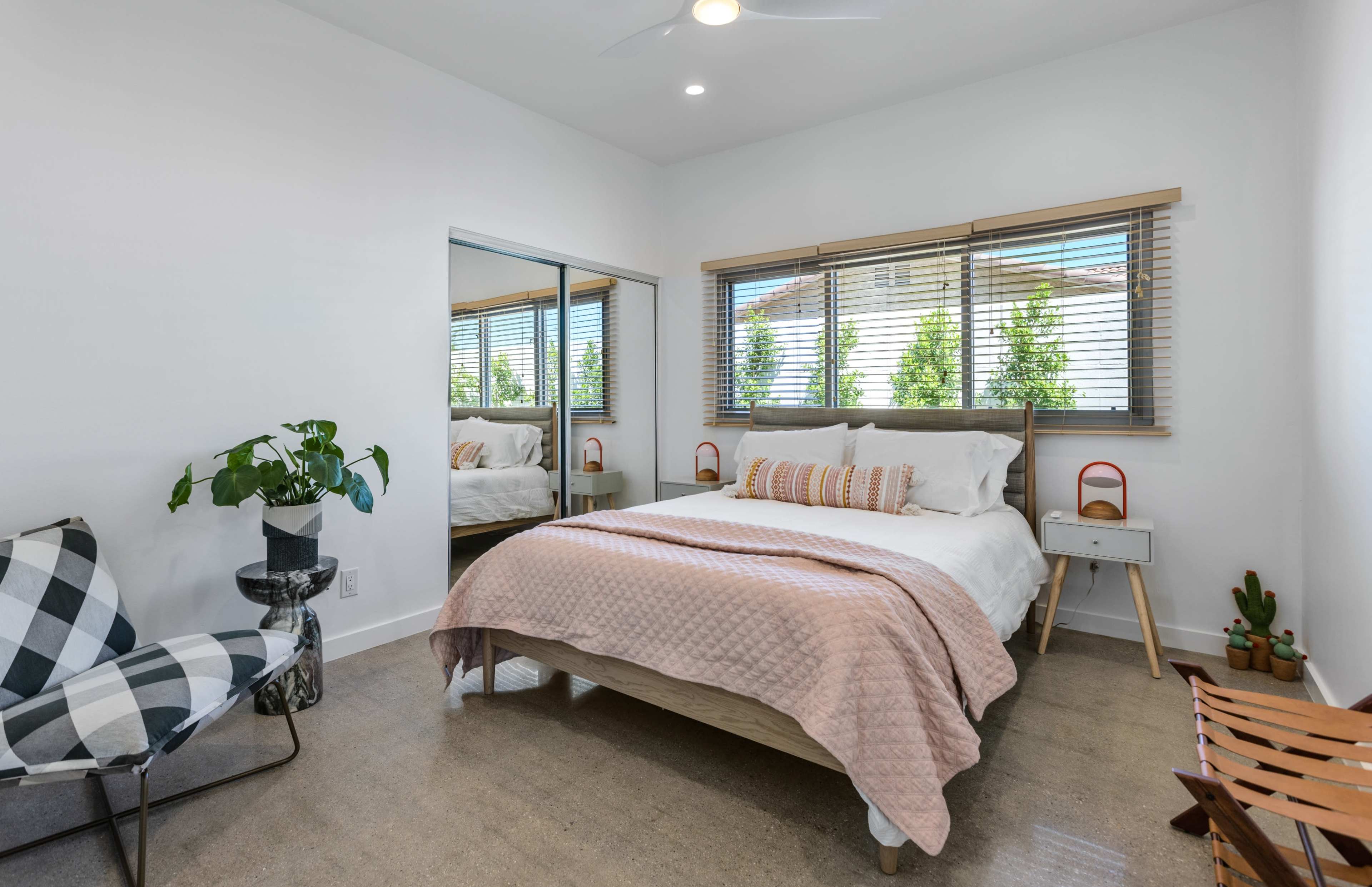 A modern bedroom features a bed with a light quilt, a mirror reflecting the space, and a chair beside a small table and potted plant.